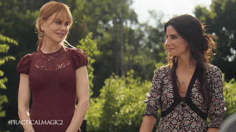 Two women stand outdoors in a lush green garden on a sunny day, smiling at each other. The woman on the left has strawberry-blonde hair pulled back and wears a burgundy lace dress. The woman on the right has long dark hair and wears a patterned dress with