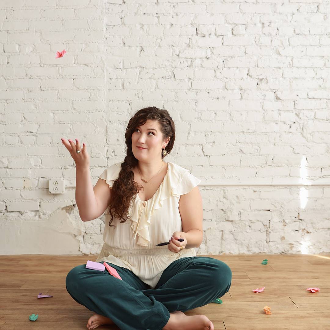 A woman sits cross-legged on a wooden floor in front of a white brick wall, smiling as she tosses colorful crumpled sticky notes into the air.