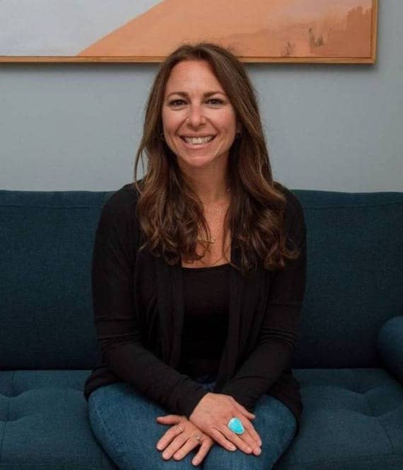 A woman with long brown hair sits on a dark blue couch smiling at the camera