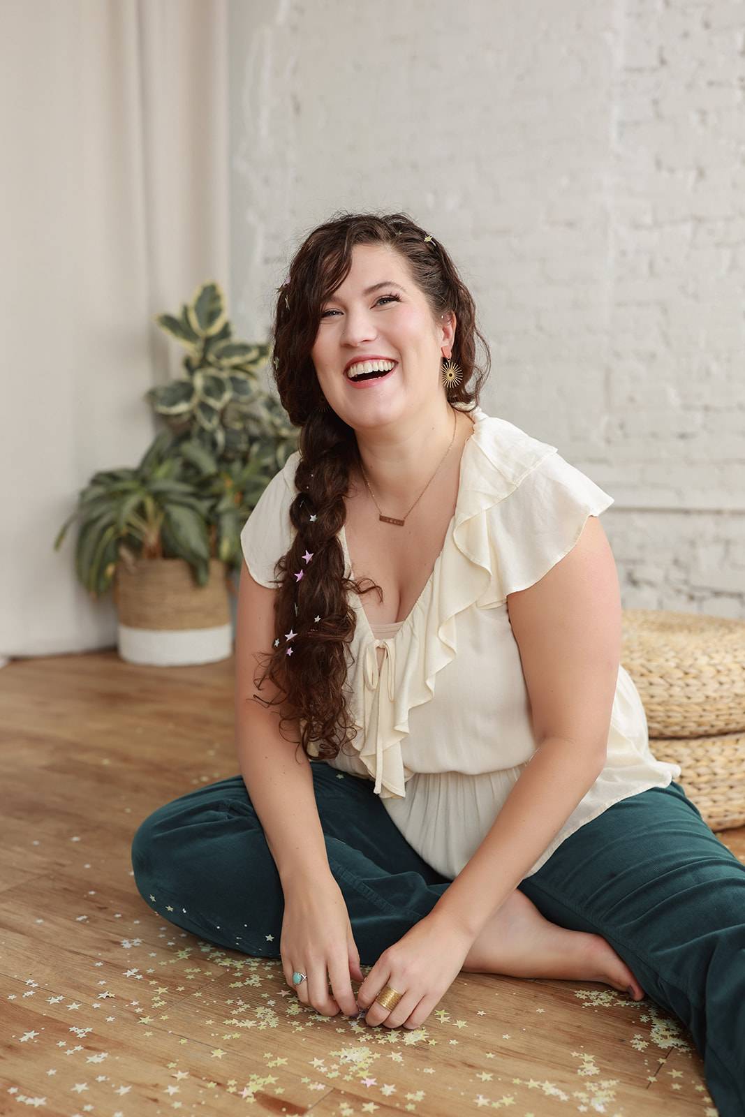 A smiling woman with long brown wavy hair in a side braid sits cross-legged on a wooden floor. She wears a cream ruffled blouse and denim pants, with confetti scattered around her. A plant and woven pouf sit in the background against a white brick wall.