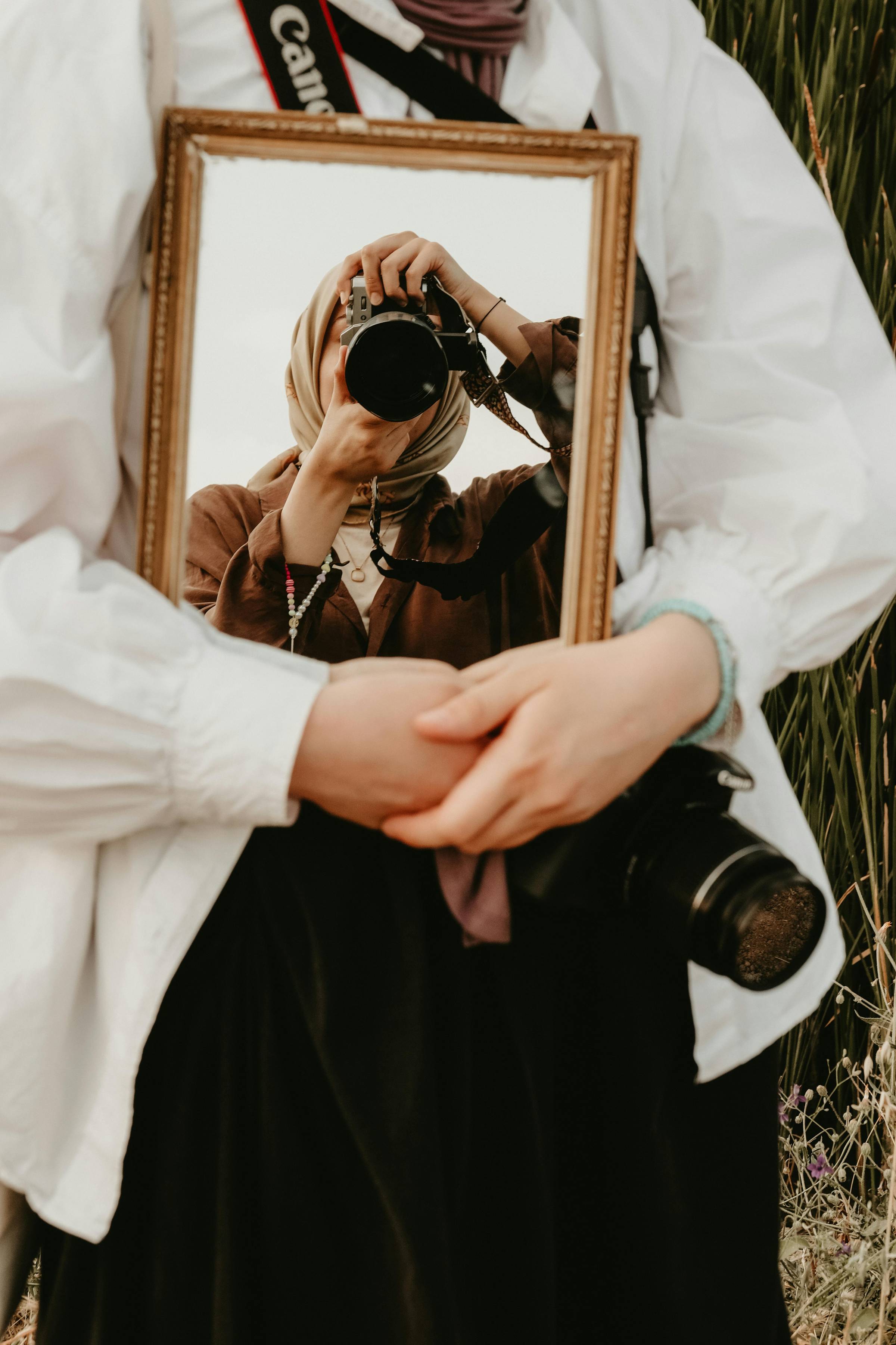 Mirror reflection of a photographer holding a camera while standing beside another person in soft, natural light.