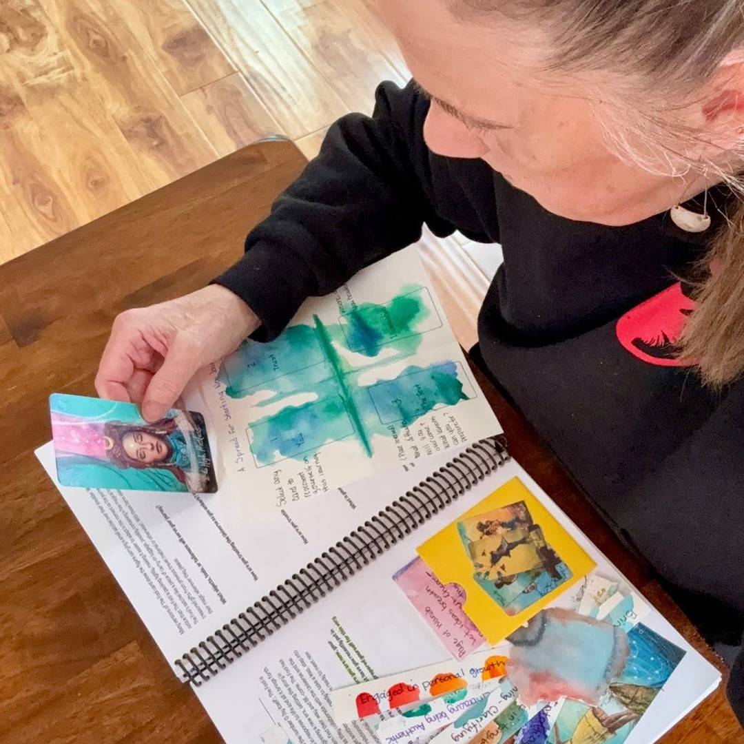 A person sits at a wooden table engaging with the Tarot for Creative Spirits workbook. They're holding a Tarot card featuring a serene woman while reading and writing notes on watercolor-painted pages filled with creative prompts and card reflections.