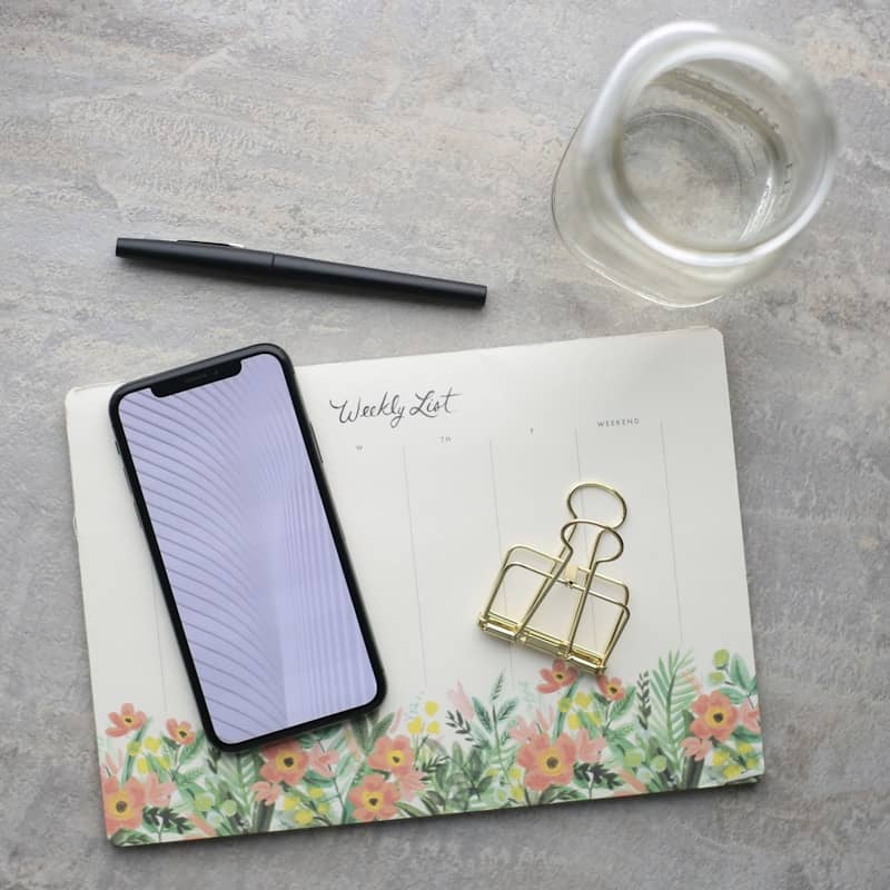 A black smartphone with a subtle wavy pattern on its screen rests on a floral-themed weekly planner. Beside it, there is a black pen, two gold binder clips, and a clear glass carafe set against a light gray tabletop.