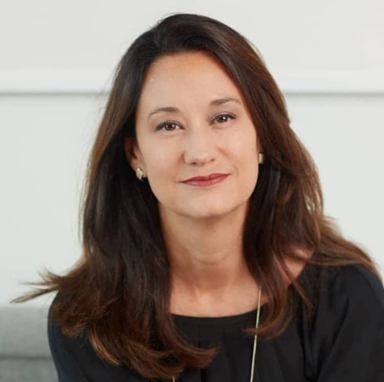 A professional headshot of a woman with shoulder-length brown hair, wearing a black blouse and subtle earrings, facing the camera with a gentle smile against a light-colored background.