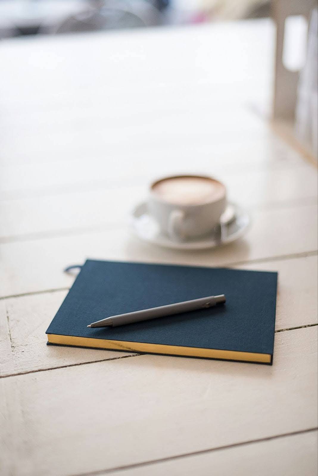 A cozy flat lay featuring a dark blue notebook with a pen resting on top, placed on a white wooden surface. In the background, there is a blurred cup of coffee in a white saucer, adding a warm ambiance.