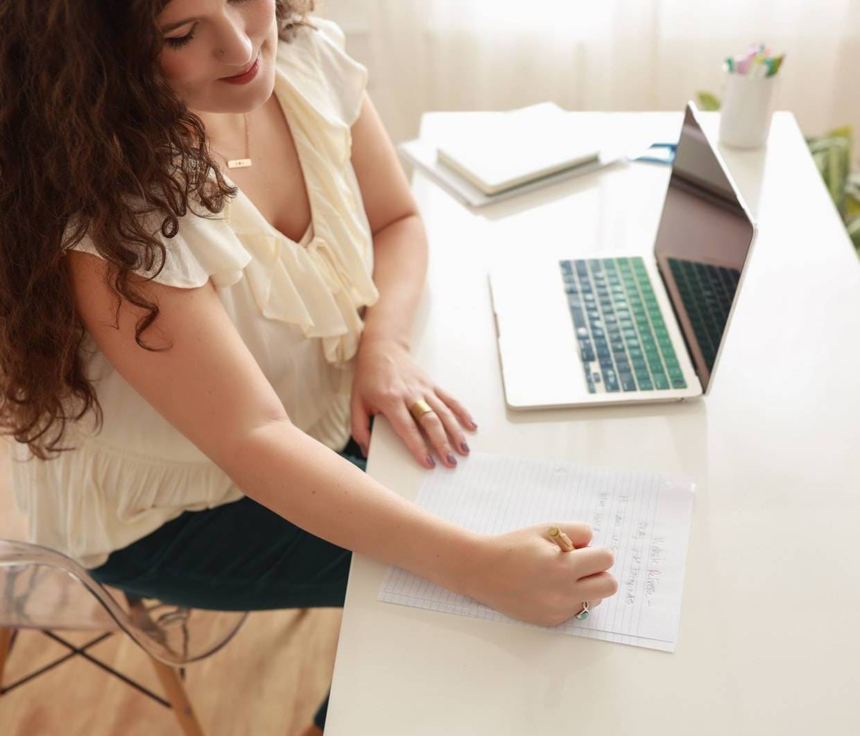 A woman with long curly hair sits at a white desk, writing in a notebook beside an open laptop and closed books.