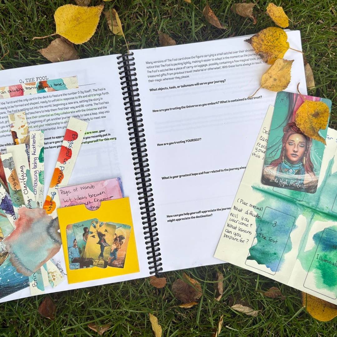 An open spiral-bound Tarot for Creative Spirits workbook rests on grass among yellow leaves. The pages are decorated with Tarot cards, watercolor art, and handwritten reflections. The visible section focuses on “The Fool” card and includes creative journaling prompts.