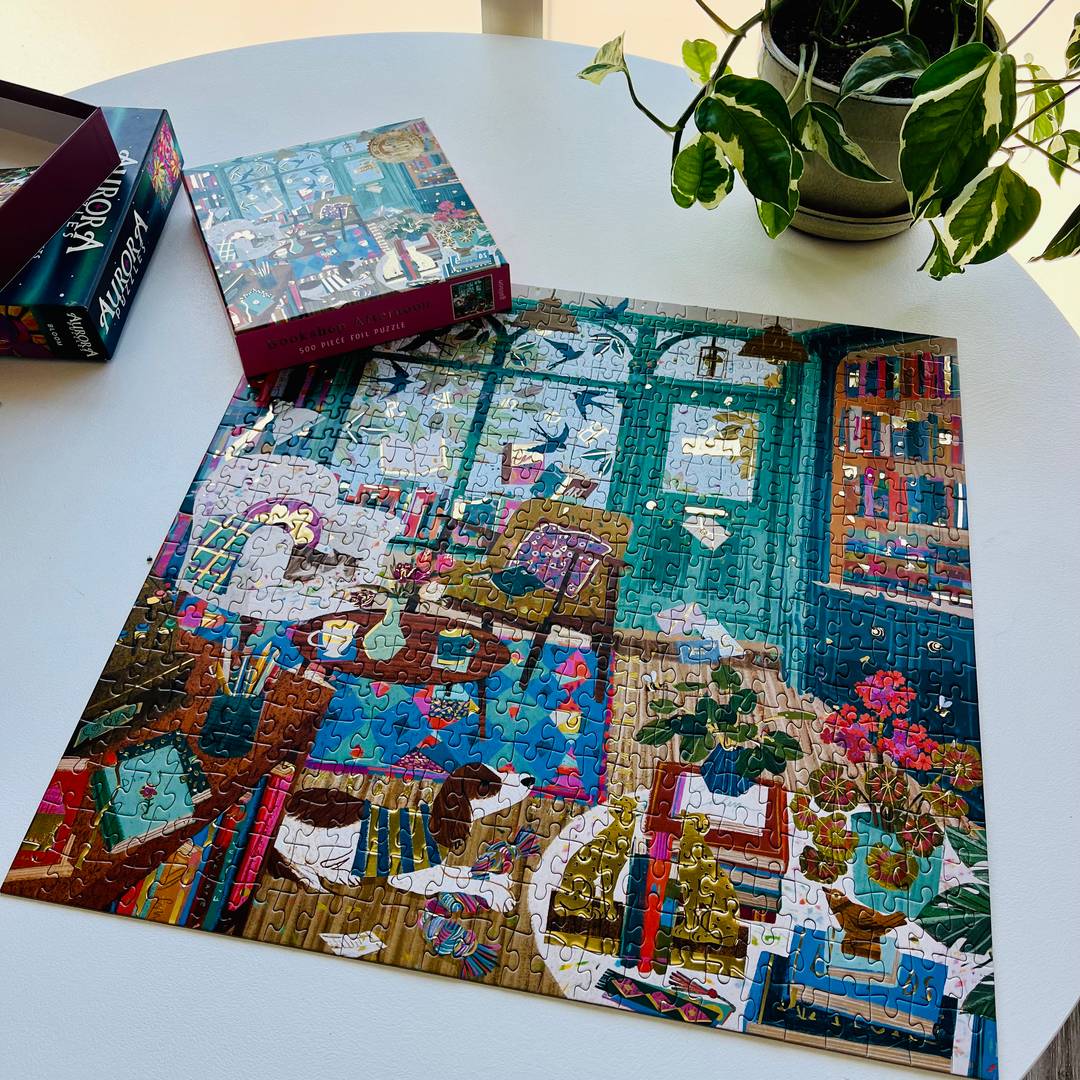 A partially completed colorful jigsaw puzzle spread across a round white table, with the puzzle box nearby and a trailing houseplant in the corner.
