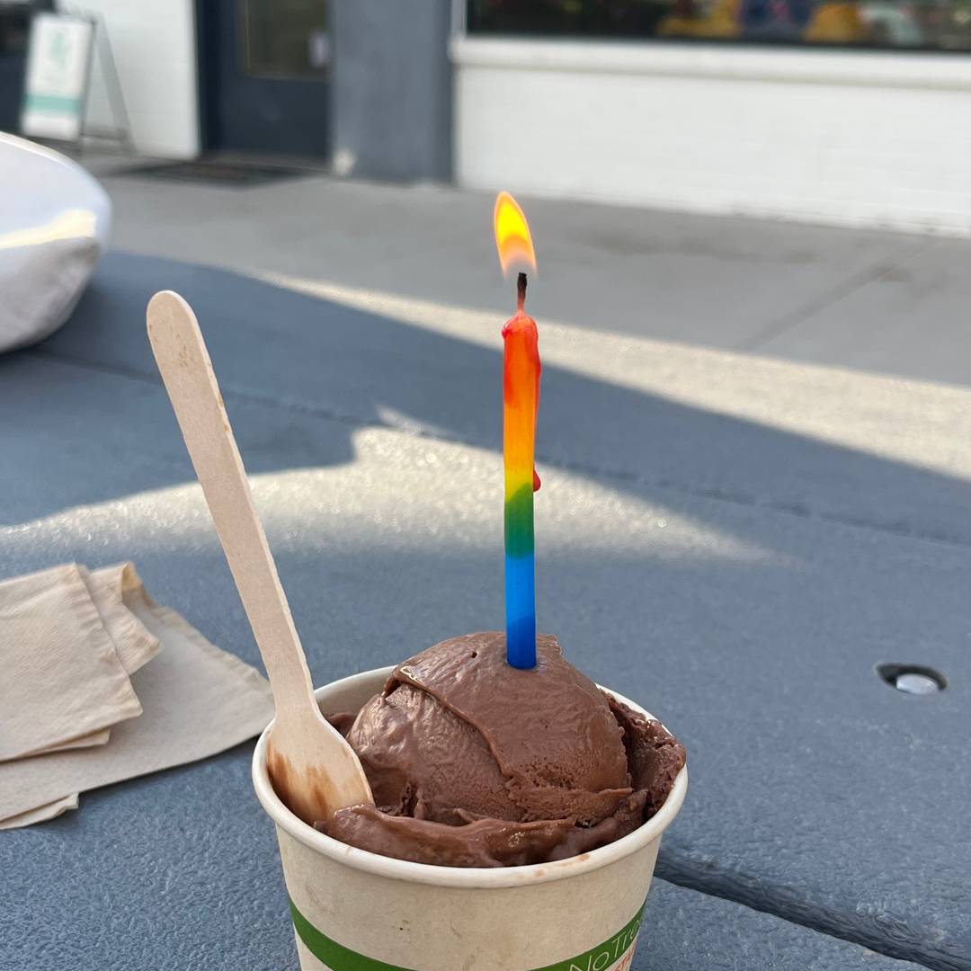 A cup of chocolate ice cream on an outdoor table with a lit rainbow birthday candle stuck in the center and a small wooden spoon resting against the cup.