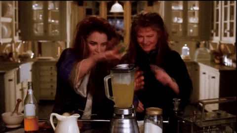 Two women with long dark wavy hair laugh in a warm, cozy farmhouse kitchen with glass-front cabinets. One pours a pale yellow drink from a blender while the other dances beside her. A pitcher, a jar, and a bottle sit on the counter in front of them.