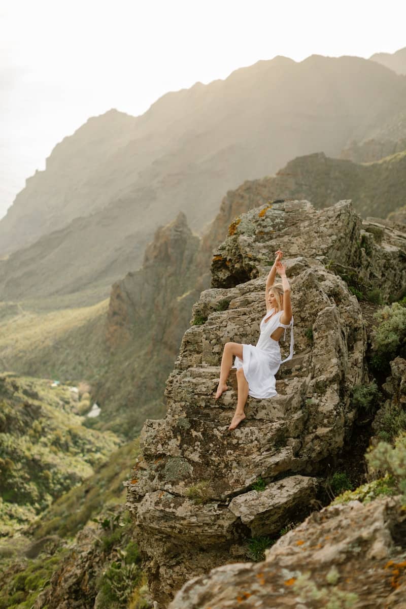 Woman in white dress poses on rocky mountain overlooking valley.