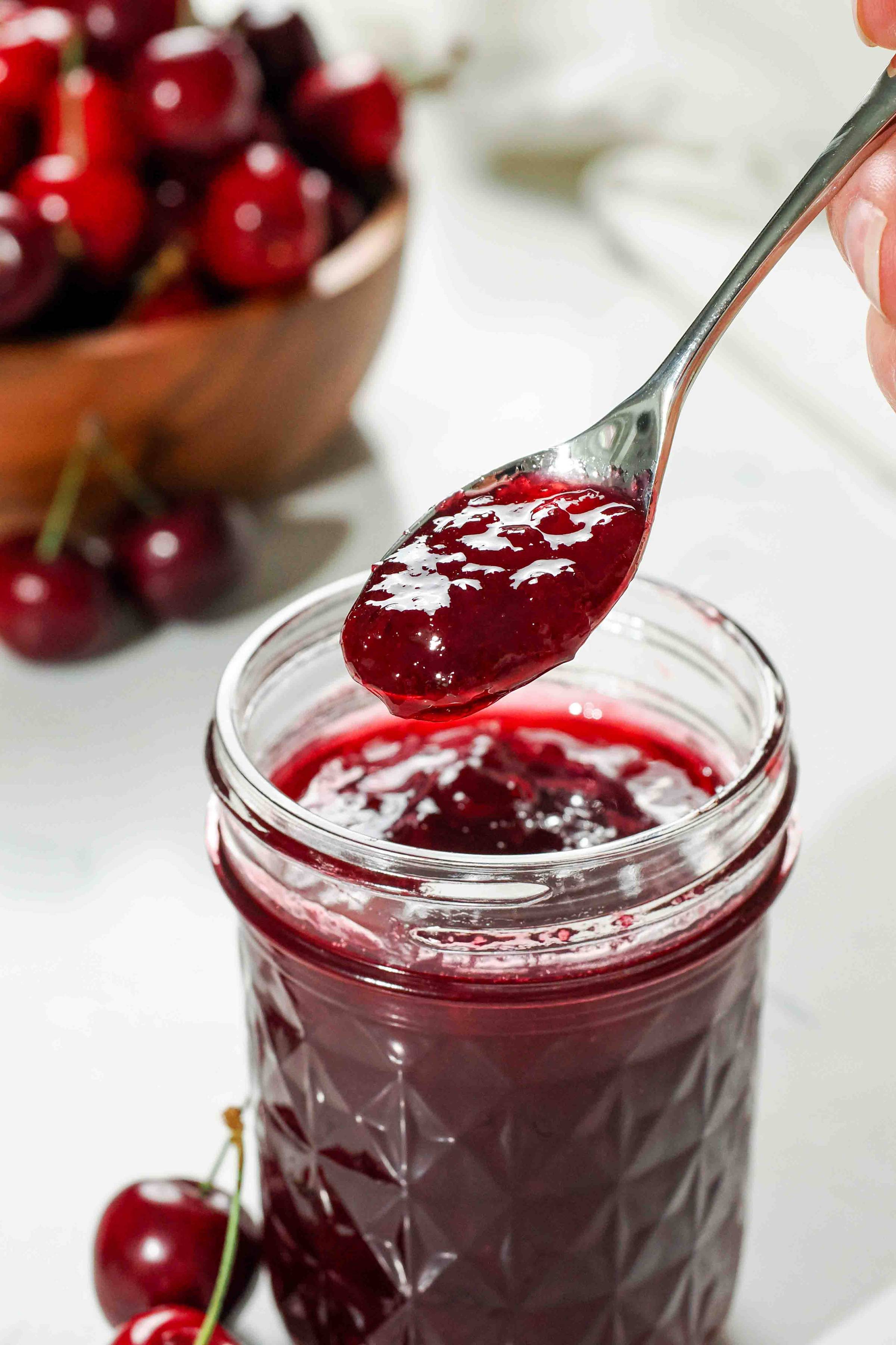 A spoon holds up cherry jam from a jar