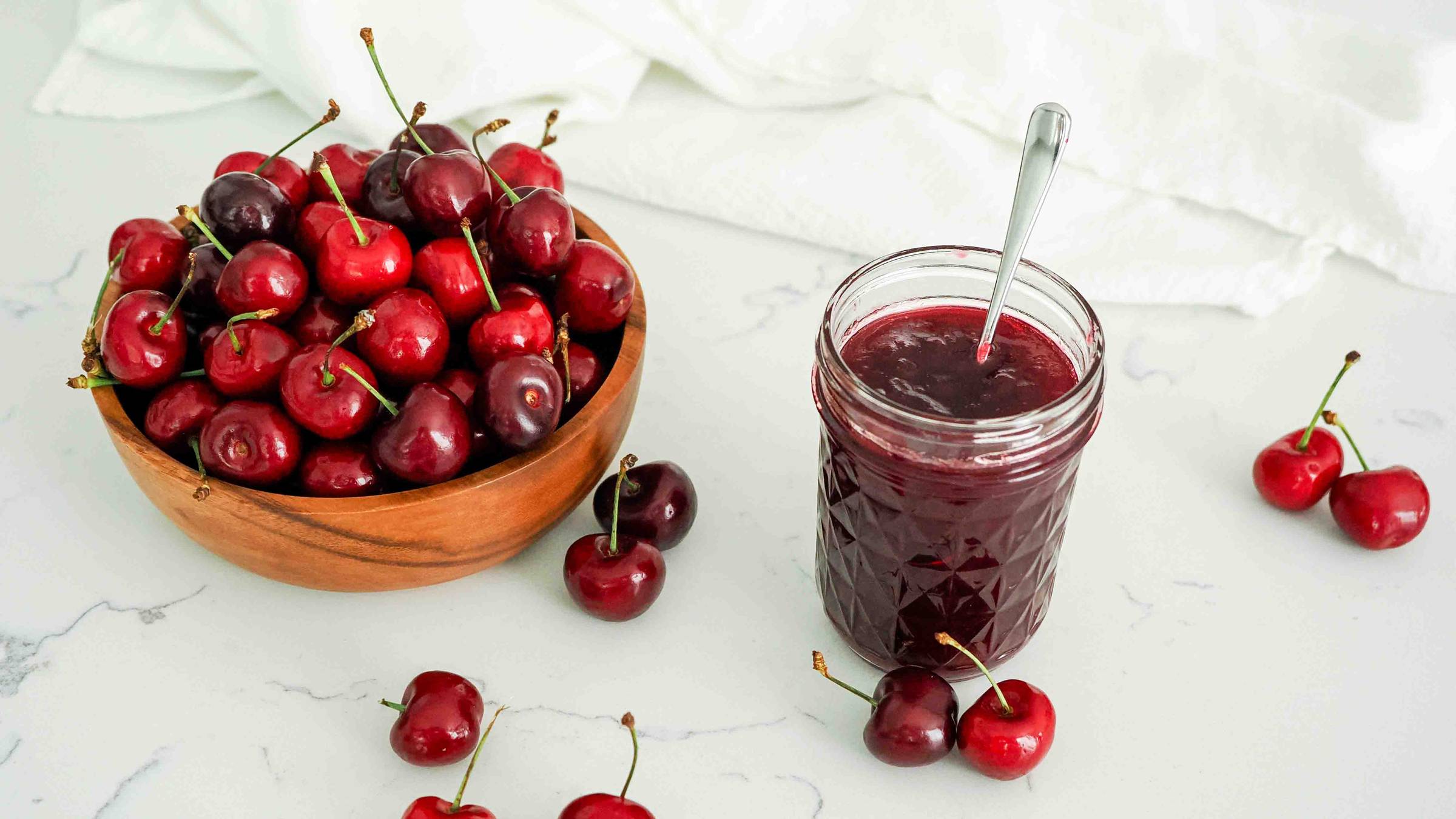 A jar of cherry jam with a bowl of cherries.