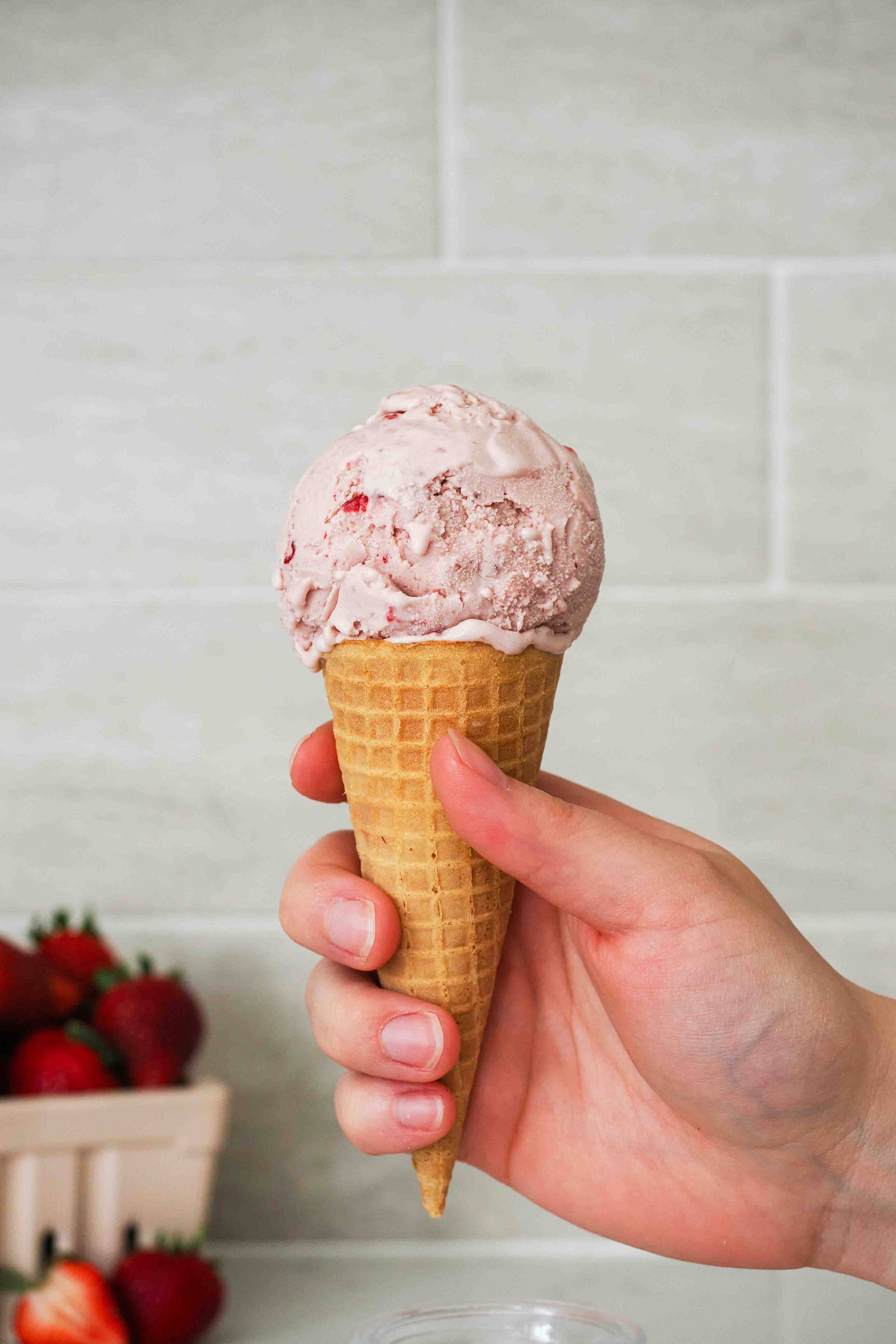 A hand holds up an ice cream cone with a scoop of strawberry ice cream.