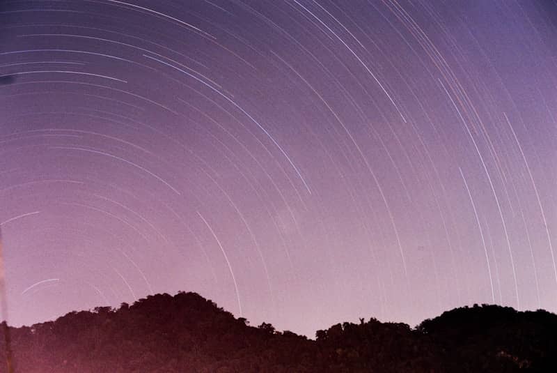 Star trails arc across a purple night sky over mountains.