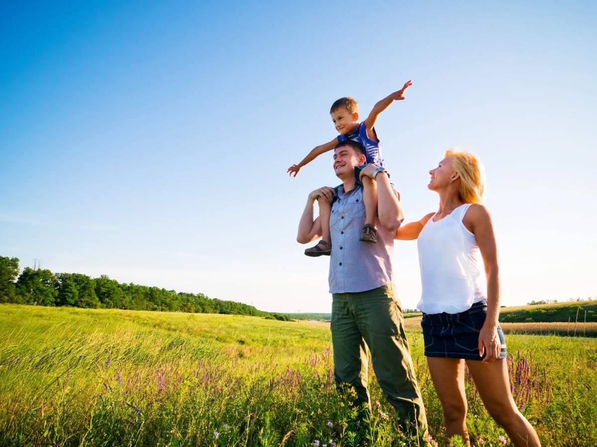 A man carries a child on his shoulders in a grassy field under a clear blue sky.