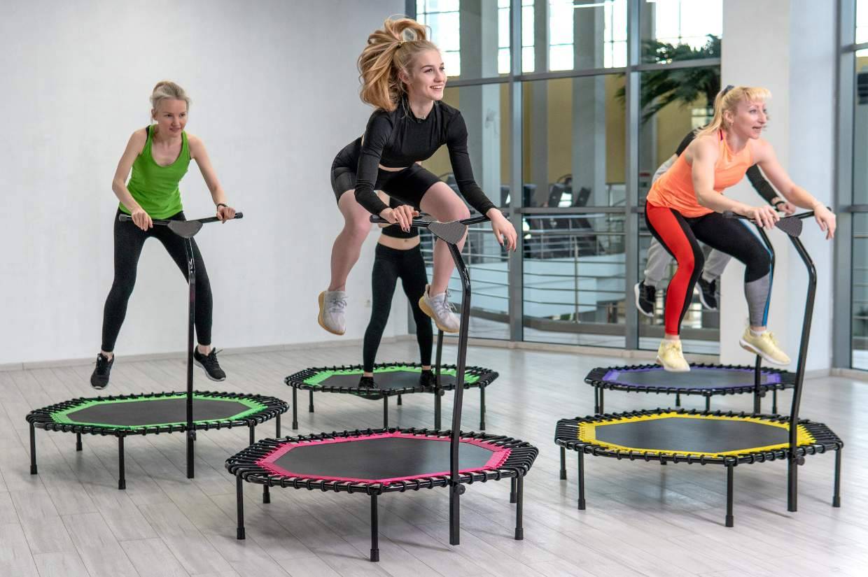 Four women are exercising on mini trampolines with handlebars in a brightly lit room. 