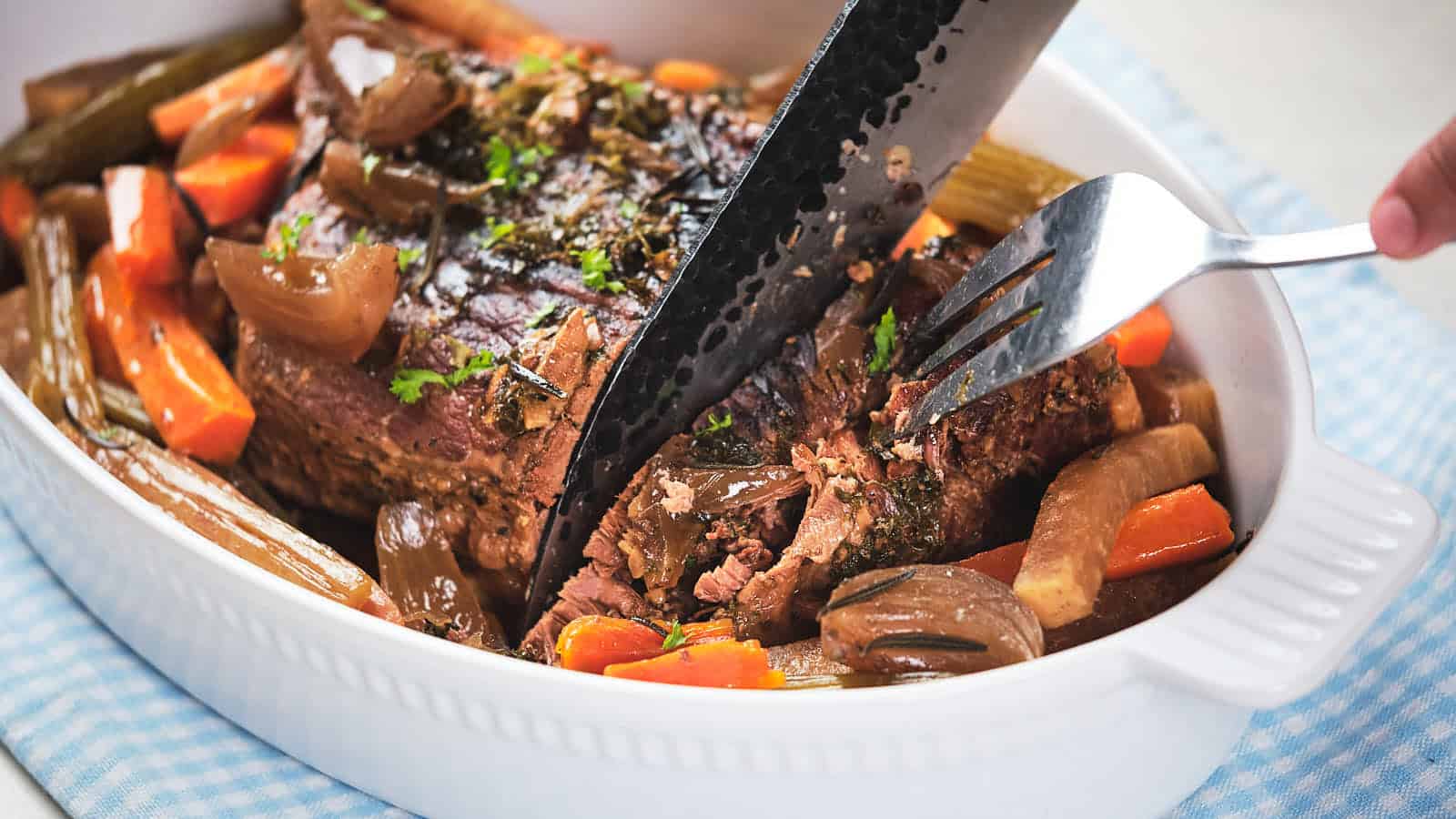 A cooked pot roast in a white oval casserole dish, being cut with a pair of black kitchen shears.