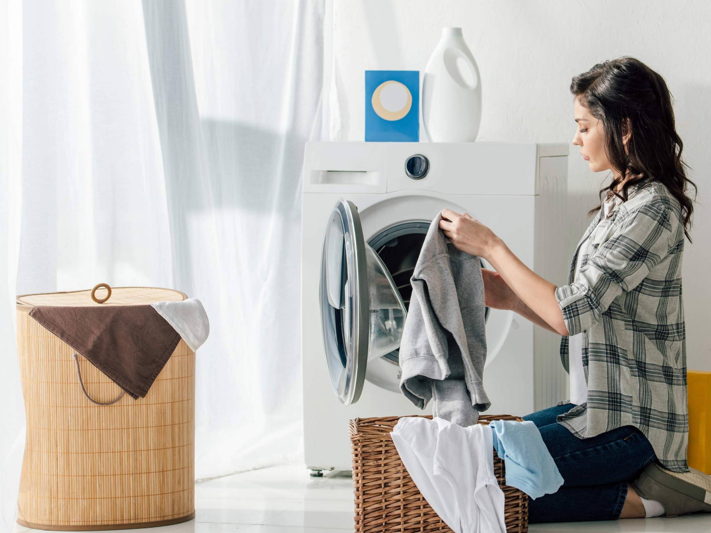 A woman kneeling on the floor loads clothes into a front-loading washing machine.