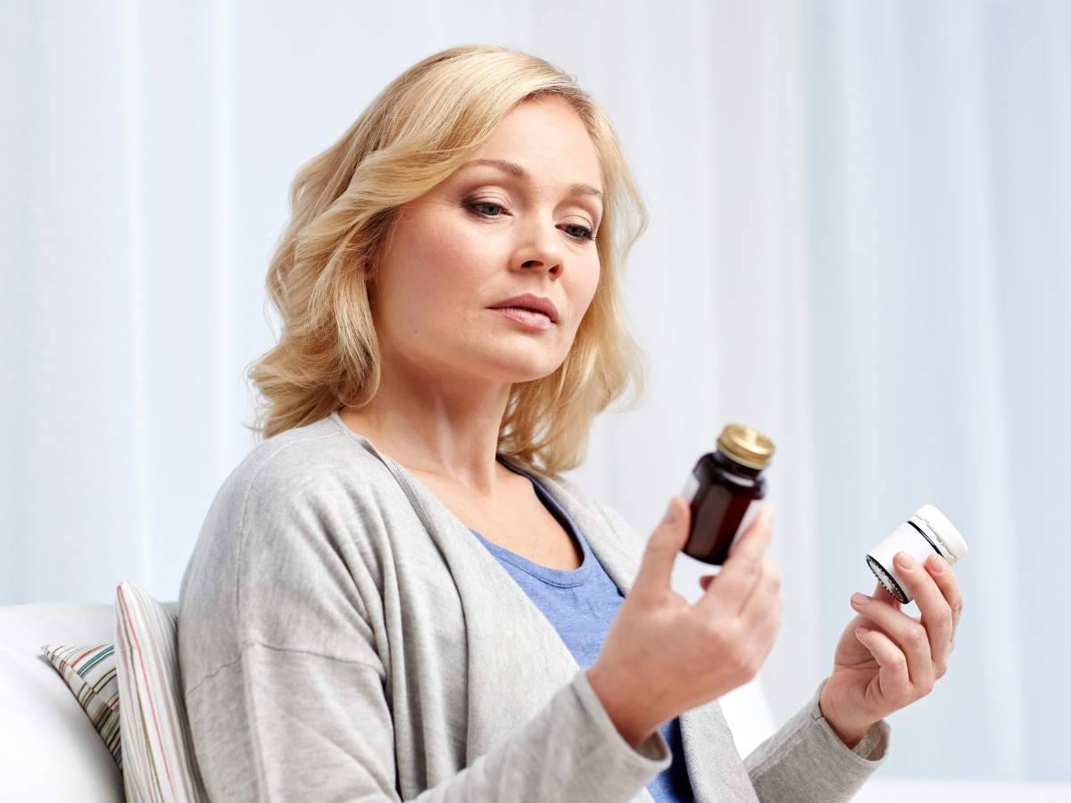 A woman with blonde hair examines a bottle of medication in one hand and holds another container in the other hand while seated indoors.