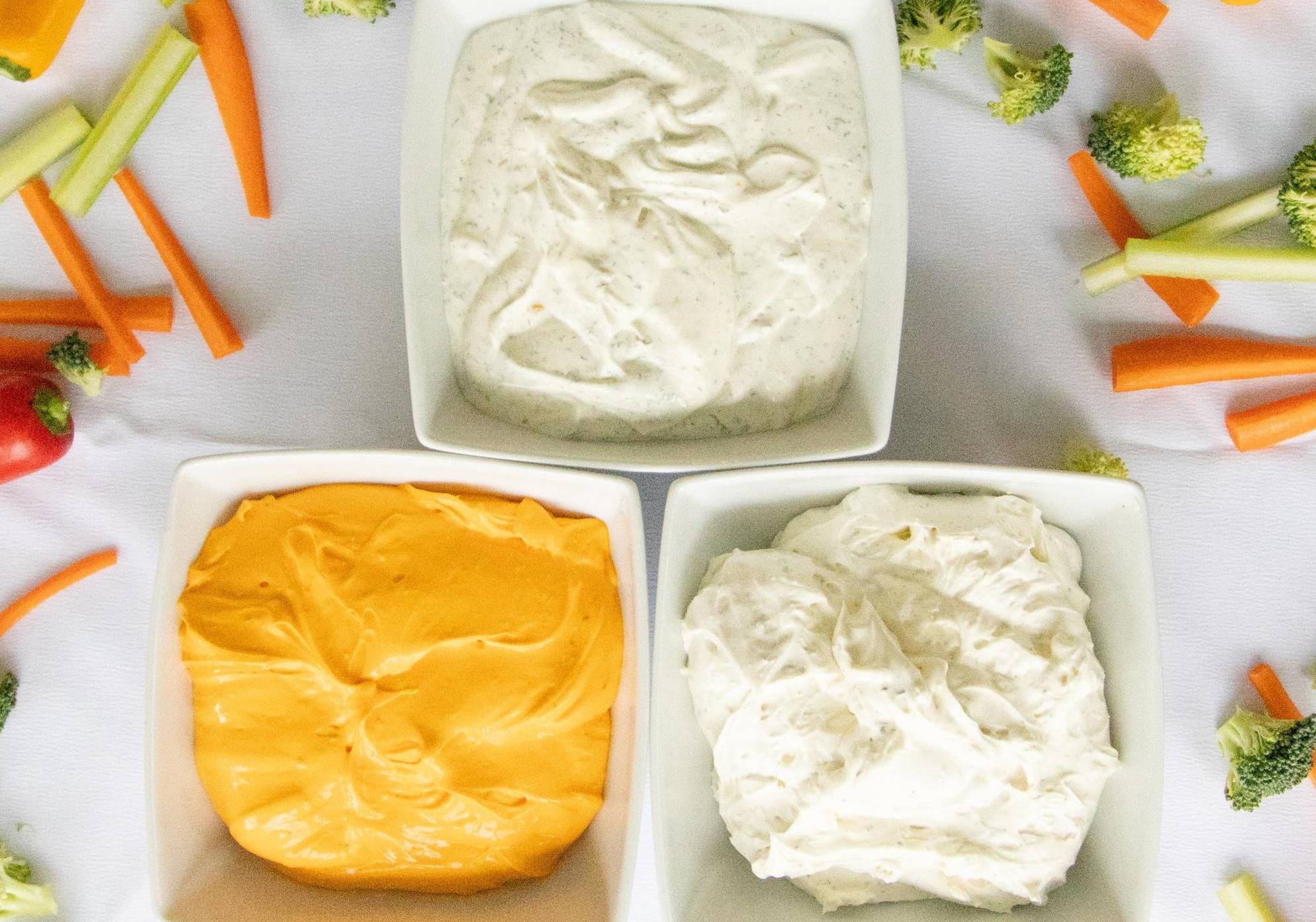 3 bowls of dip on the counter with veggies laying nearby