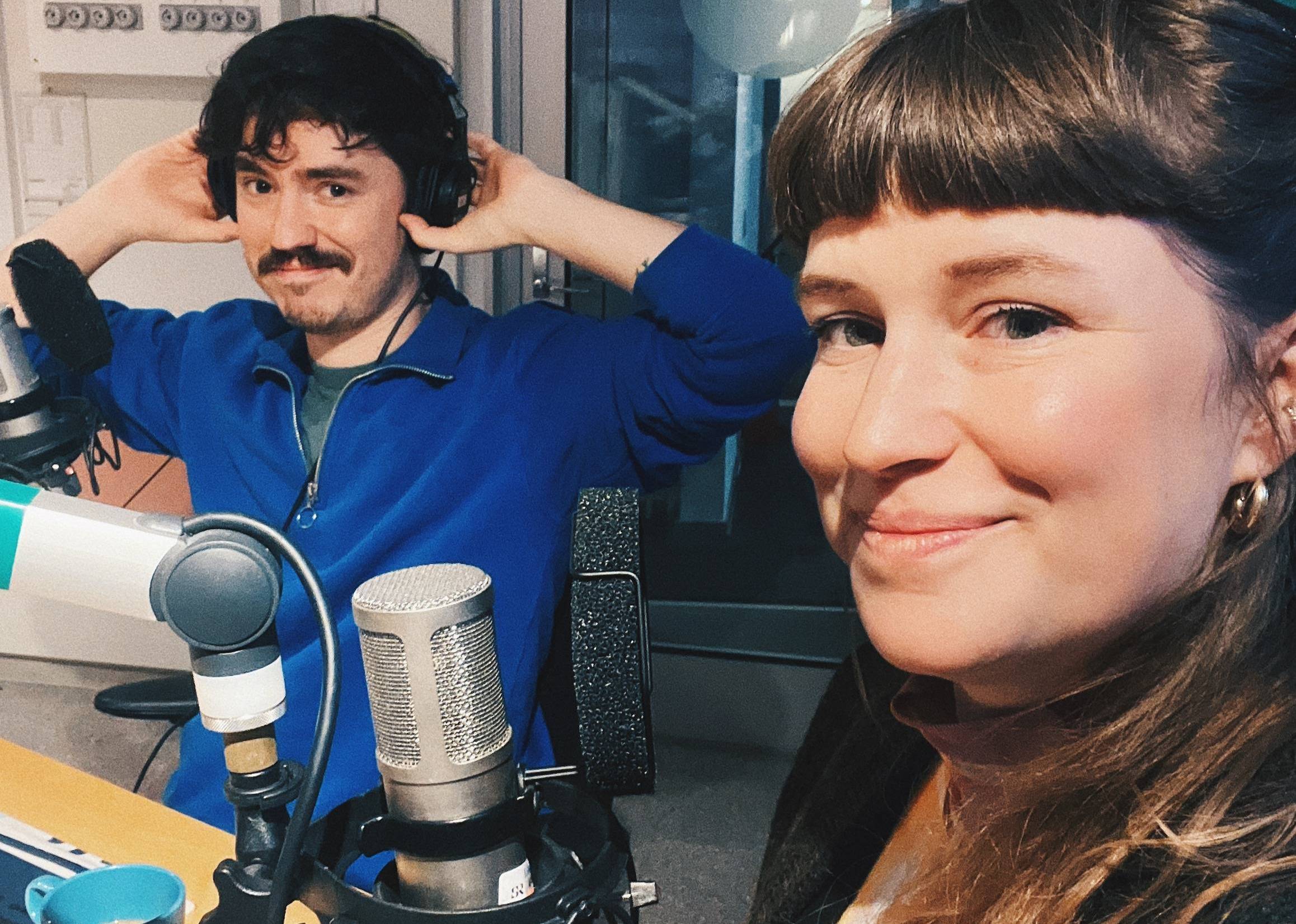 Elin and Mario sitting in front of two microphones, with headphones on, looking excited and secretive.