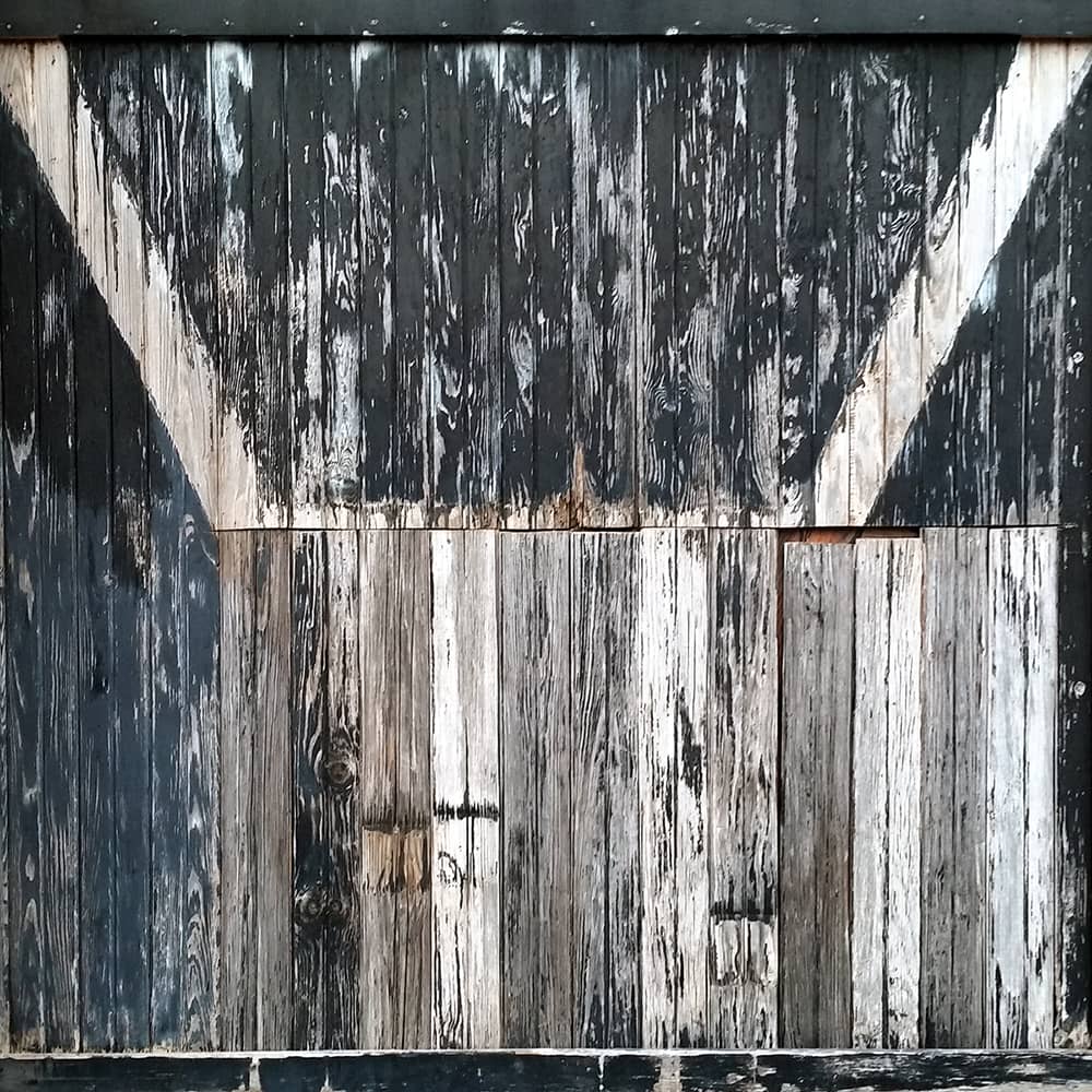 Photo of a wall made from wood slats. The black paint is fading and the wood shows signs of weathering.