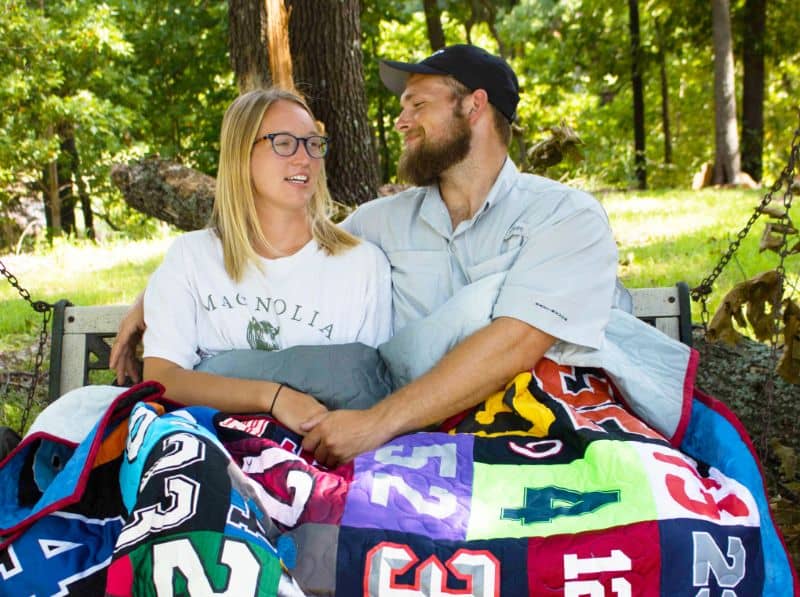 Photo of a woman and a man sitting together under a quilt made of t-shirts. They are smiling into each other's faces. Grass and woods are visible in the background.