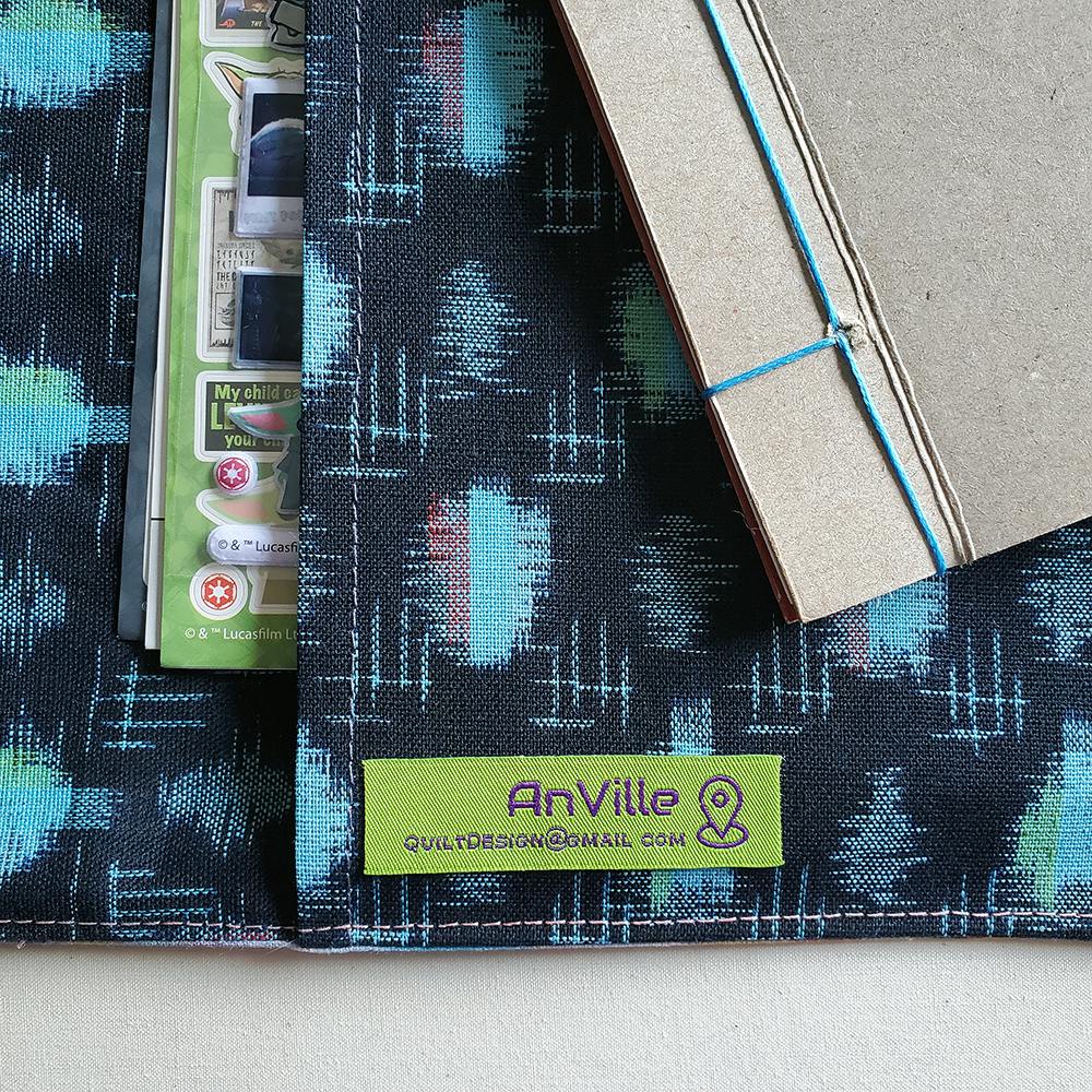 Close-up photo of a homemade cardboard sketchbook sitting on its fabric cover. A tag on the fabric reads "AnVille QuiltDesign@gmail.com"