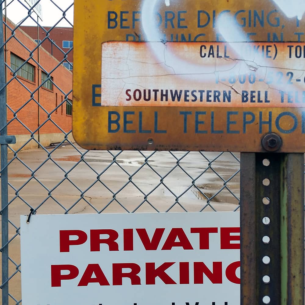 Photo of signs in front of a chain link fence, with a red brick building in the background. The signs read "Southwestern Bell Telephone" and "Private Parking."
