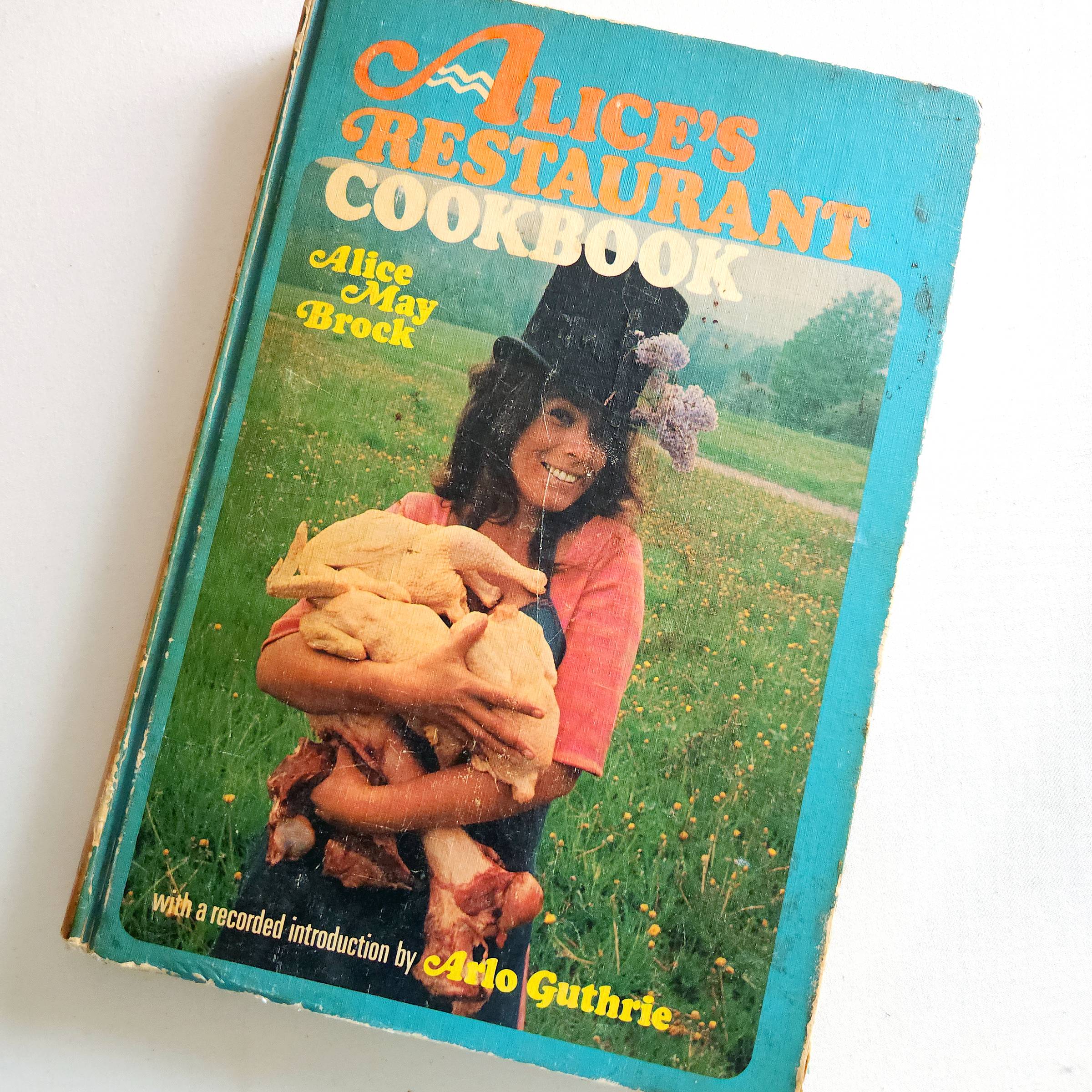 Off-kilter photo of a copy of Alice's Restaurant Cookbook by Alice May Brock published in 1969. In the cover photo, Alice is wearing a black tophat with lilac blossoms and holding several raw, plucked chickens and larger bones. Below the photo, text reads