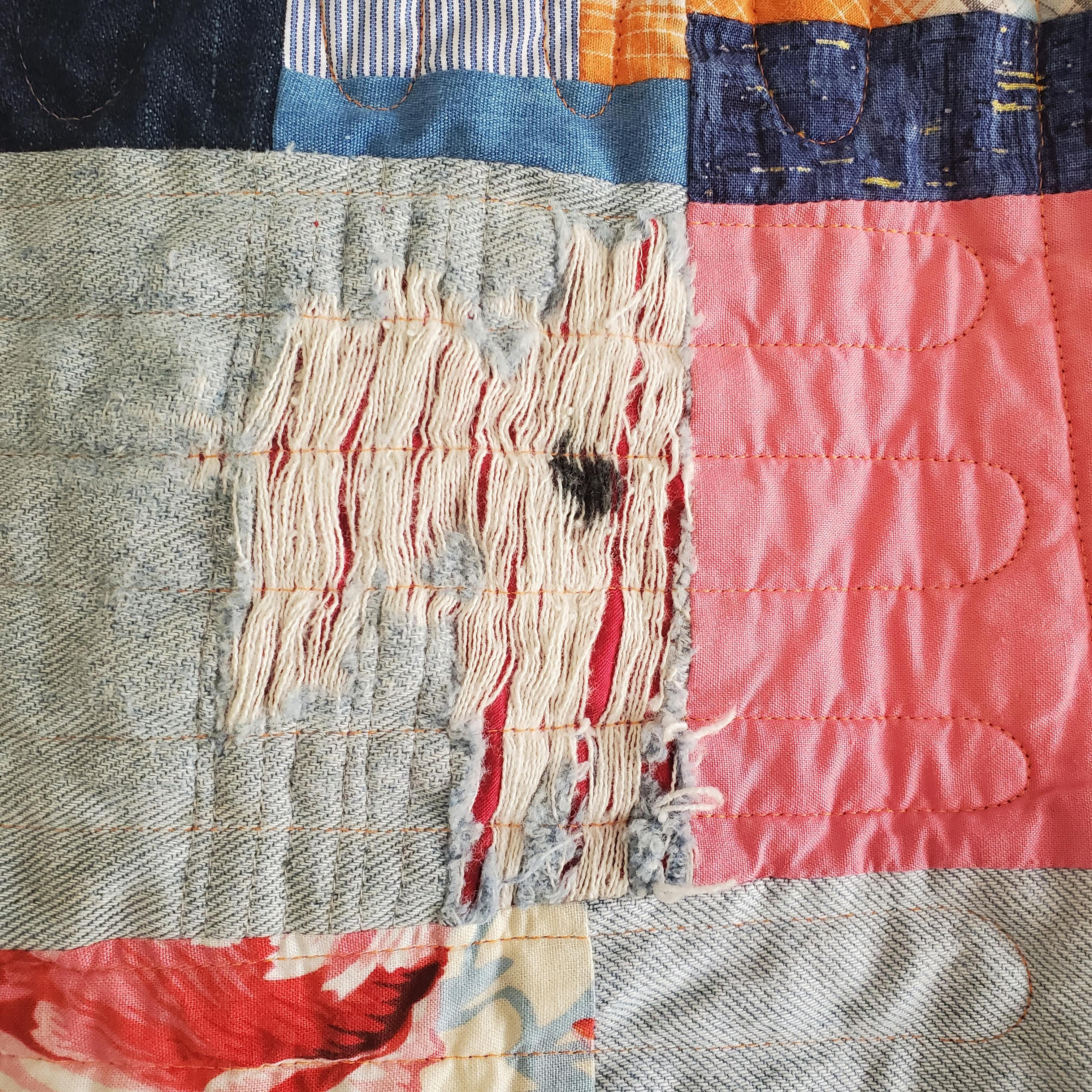 Closeup photo of a piece of worn denim sewn into a patchwork quilt. the faded blue fabric is partially disintegrated and is stained with black ink. The surrounding fabrics are red, pink, orange, and blue.