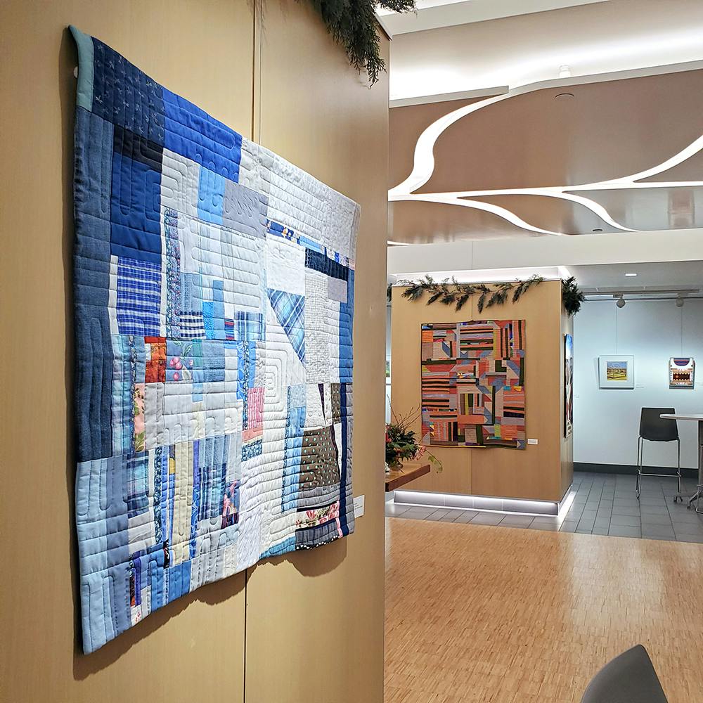 Photo of a gallery space in which quilts and other fiber art pieces are displayed on the walls.