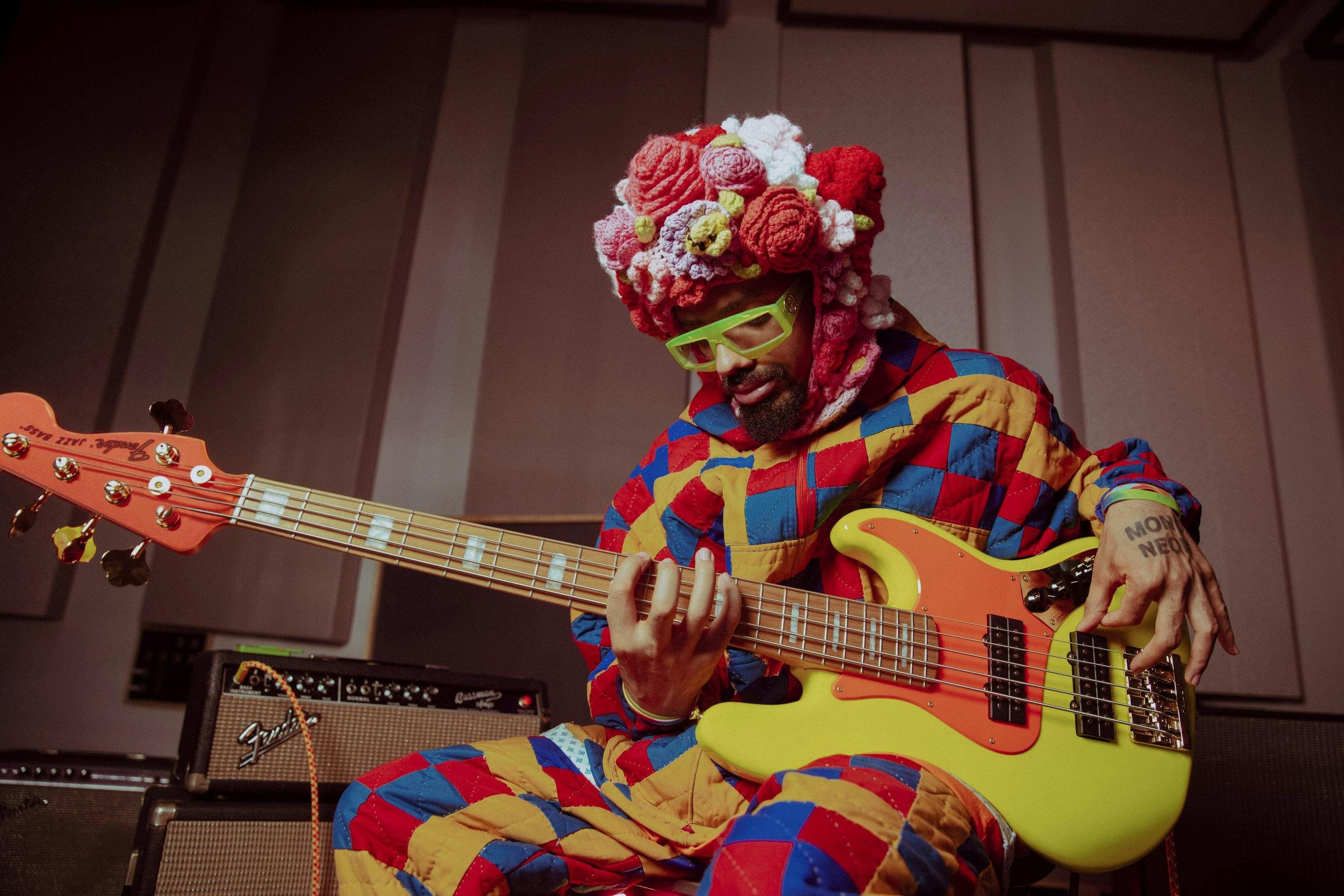 Photo of the artist MonoNeon holding his orange and yellow Fender jazz bass. He is wearing a headdress of knitted flowers, chartreuse sunglasses, and a full-body costume made from a patchwork quilt.