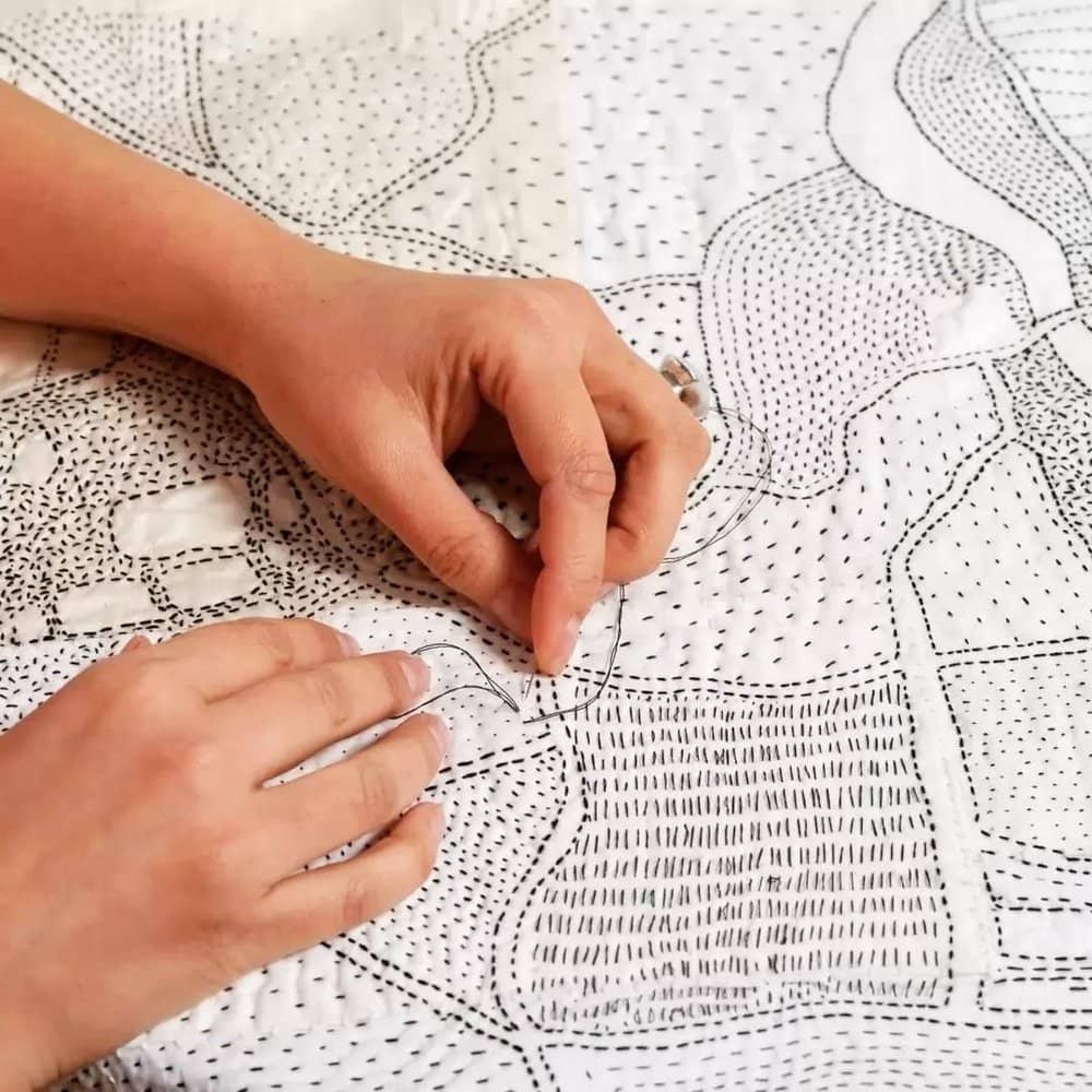 Photo of textile artist Ekta Kaul's hands stitching with black thread on a piece of white fabric.