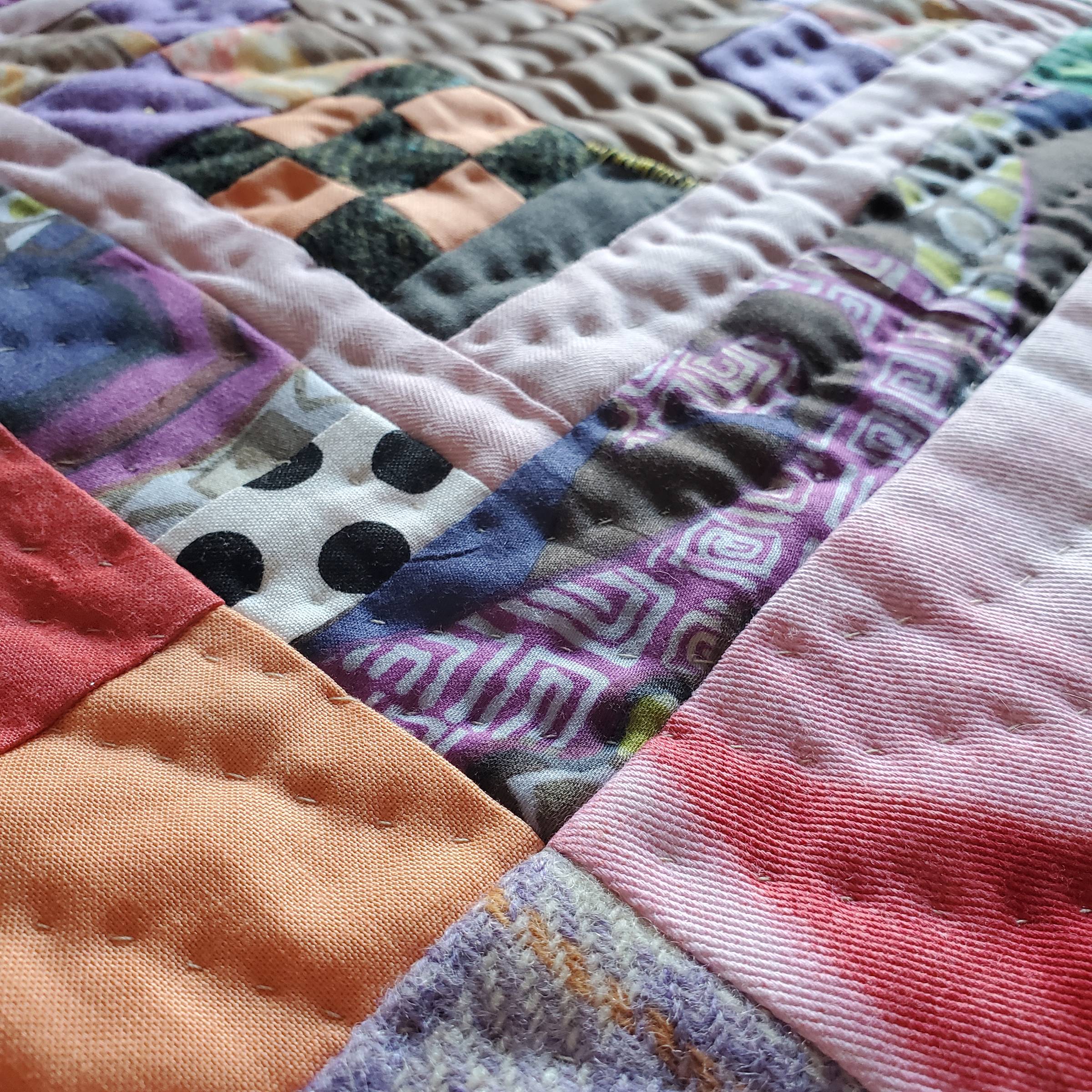 This is a detail of an improvisational patchwork quilt by Sarah Atlee. The design is a series of concentric rectangles, some subdivided into squares. The fabric colors include red, white, pink, orange, pale green, and many shades of purple. In this photo 
