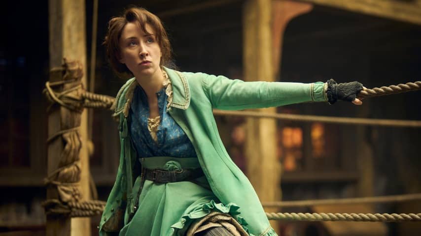 Promotional photo for the Hulu series A Thousand Blows. The image shows actress Erin Doherty as Mary Carr. She is seated in front of a boxing ring with one arm on the ropes. She has brown hair pinned up, an indignant facial expression, and a light green dress. Click image to view source.