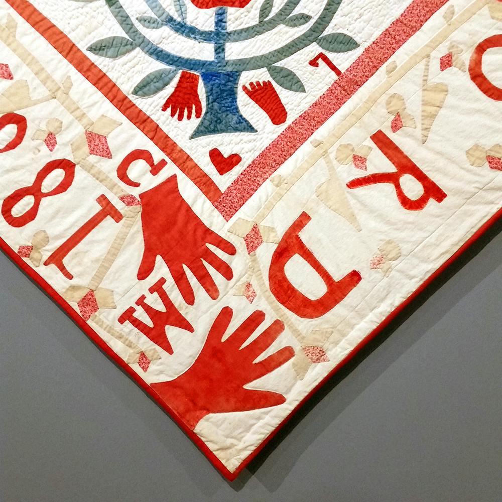 Close up photo of a 19th-century applique quilt showing hand and foot shapes in red fabric on a light background.