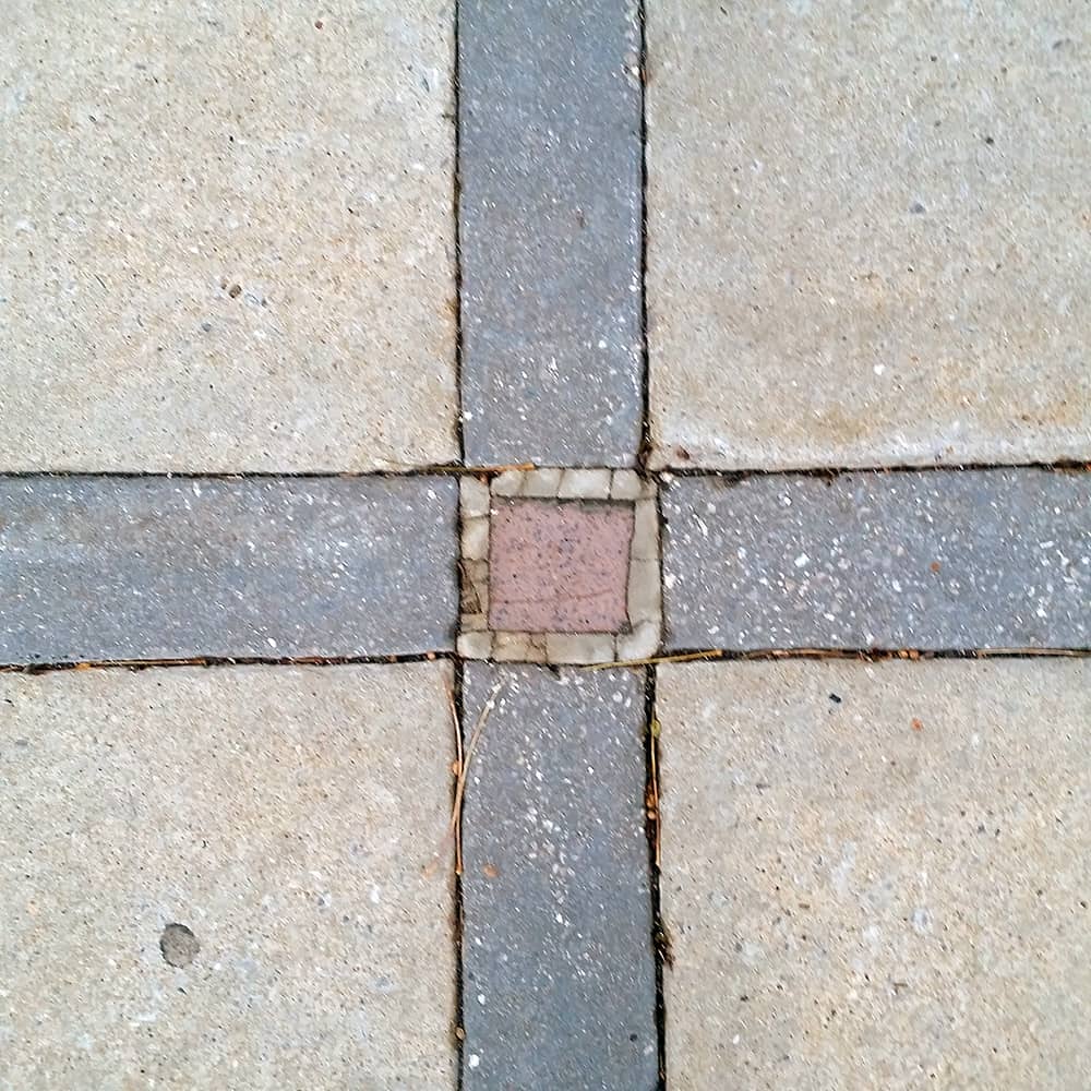 Photo of a concrete sidewalk, where four narrow bands of darker concrete meet at a red brick center.