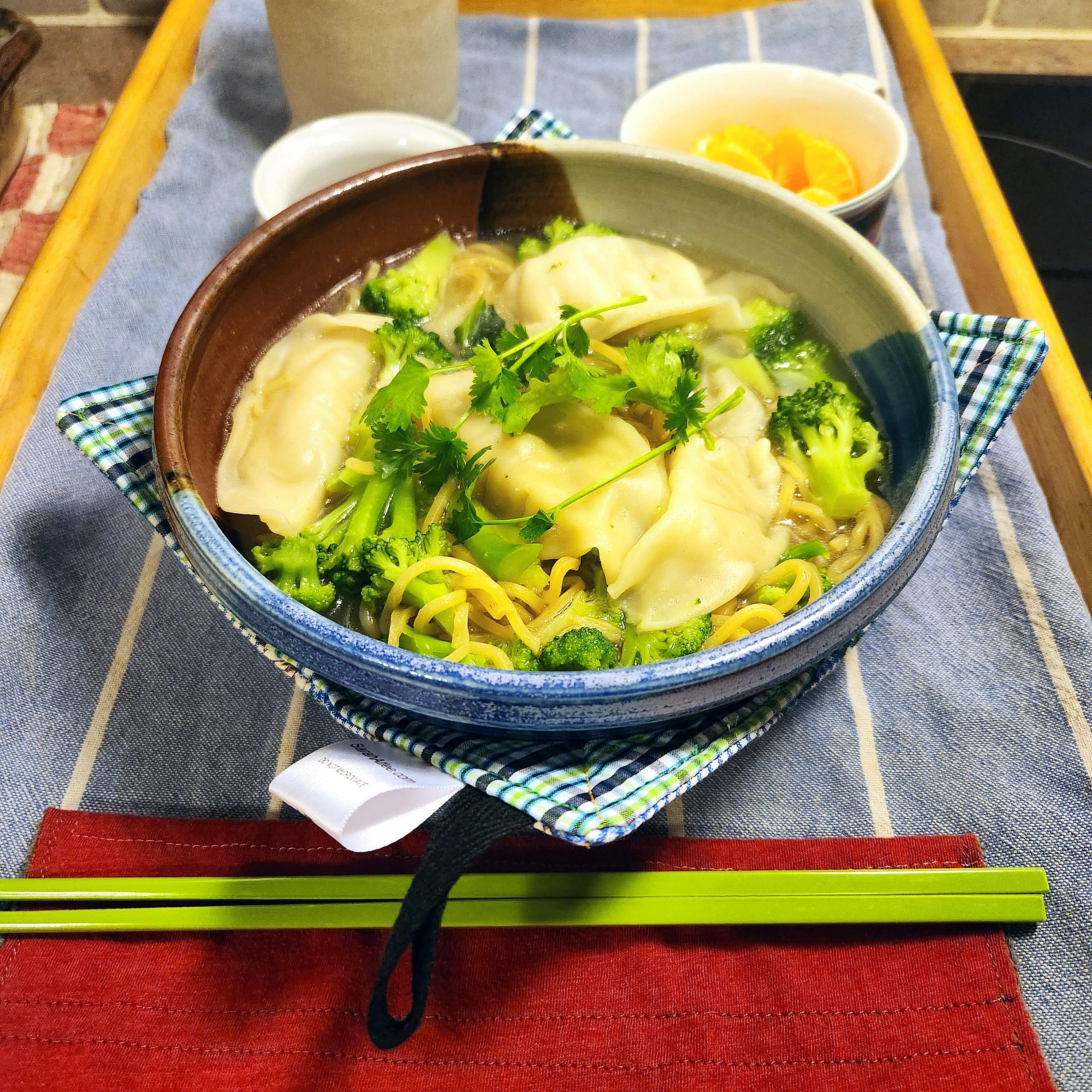 A bowl of dumpling noodle soup with veggies. The soup bowl sits inside a bowl cozy on a tray with chopsticks and other dishes. 