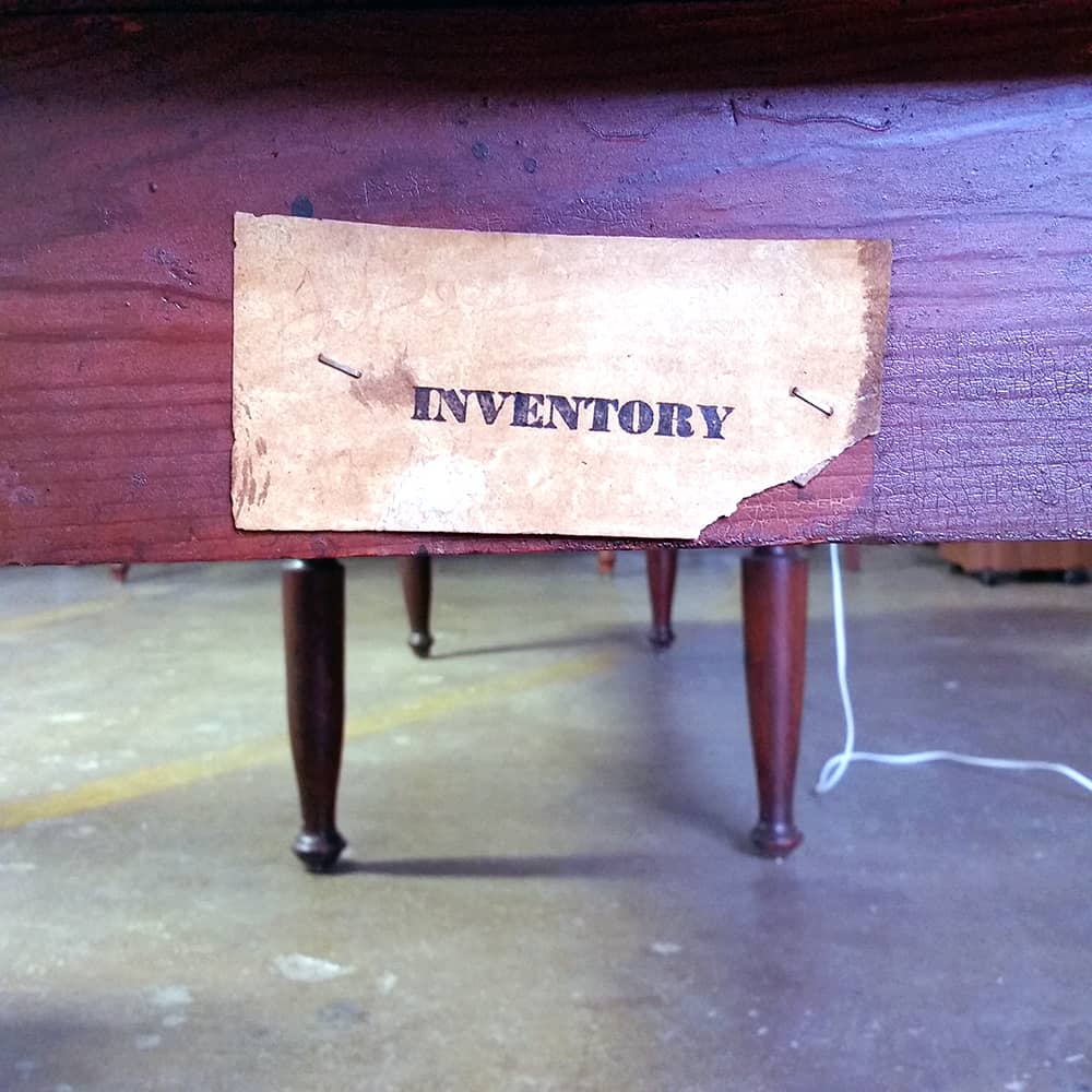 Photo of the end of a wooden table; table legs and an electrical cord are visible below. Stapled to the table is a worn paper tag that reads INVENTORY.