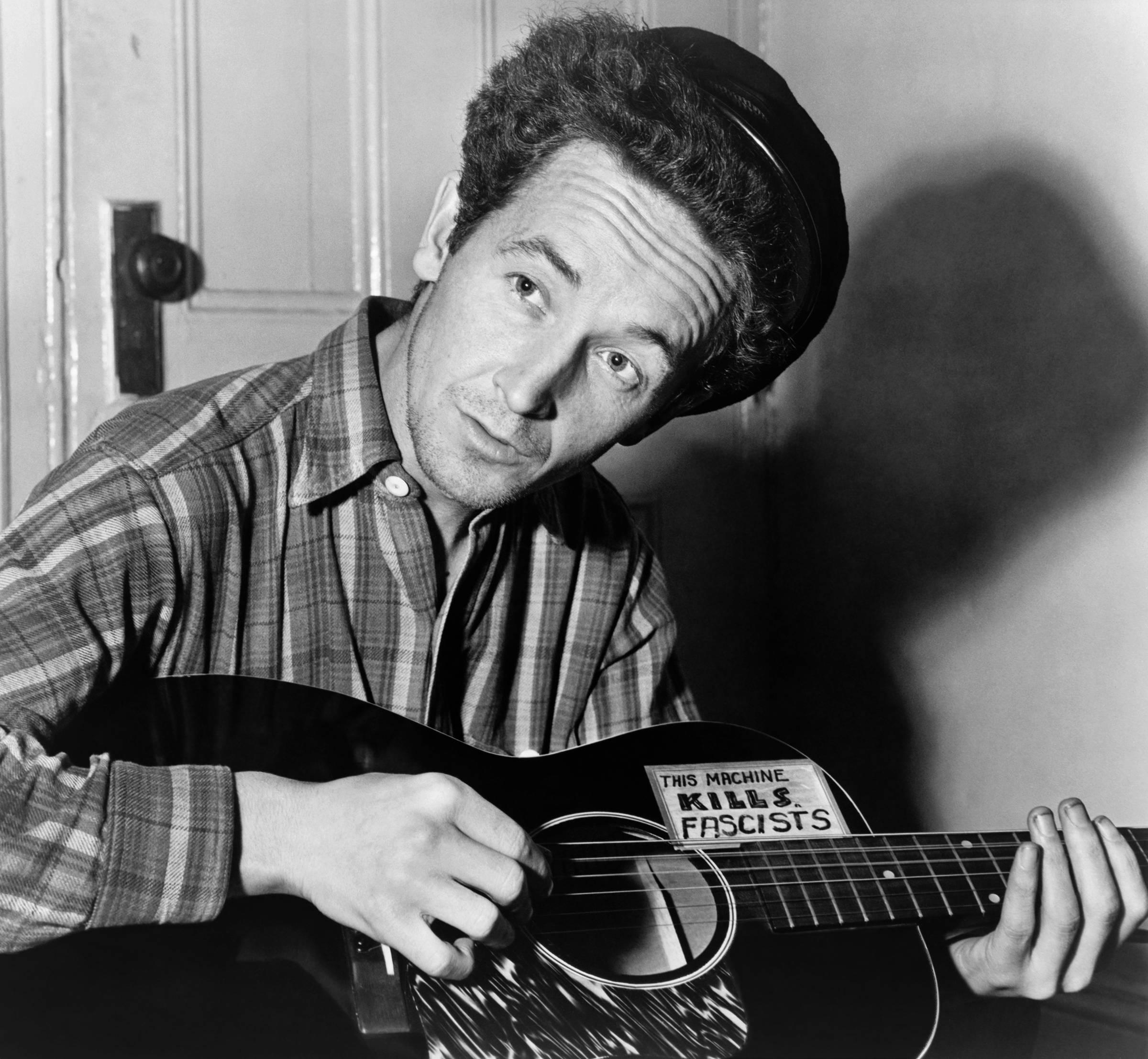 Woody Guthrie, half-length portrait, seated, facing front, playing a guitar that has a sticker attached reading: This Machine Kills Fascists