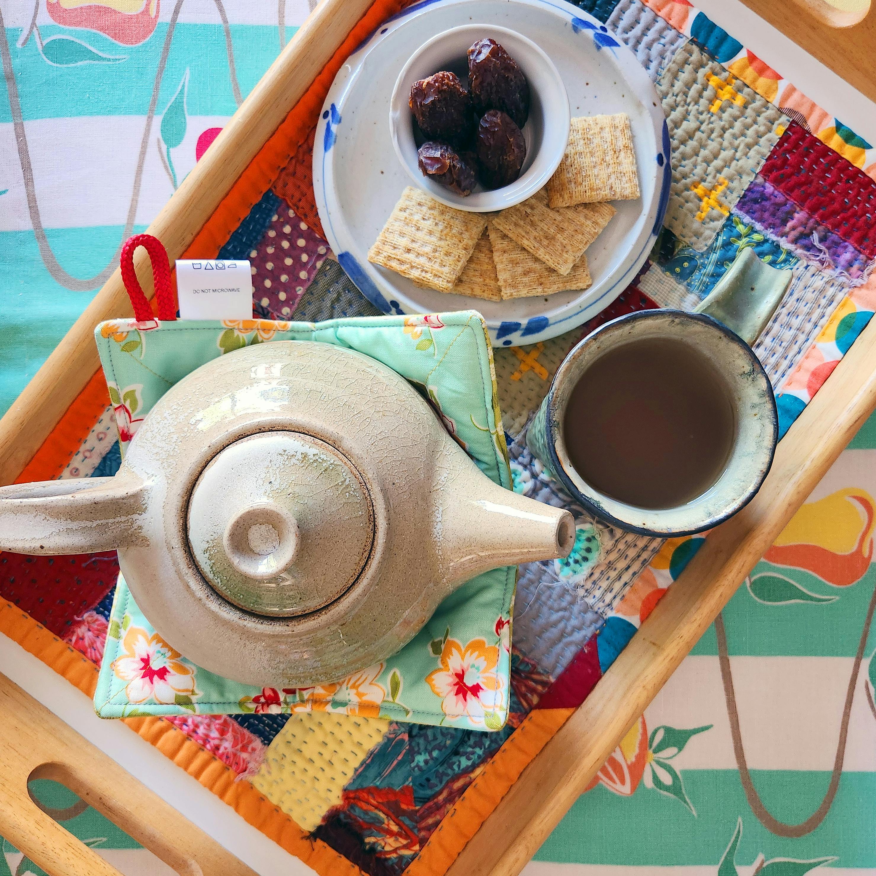 Top-down view of a tea tray including a plate with dates and crackers, a mug of tea, a ceramic teapot in a bowl cozy, and a patchwork placemat. The tray sits atop a vintage tablecloth with green and white stripes.