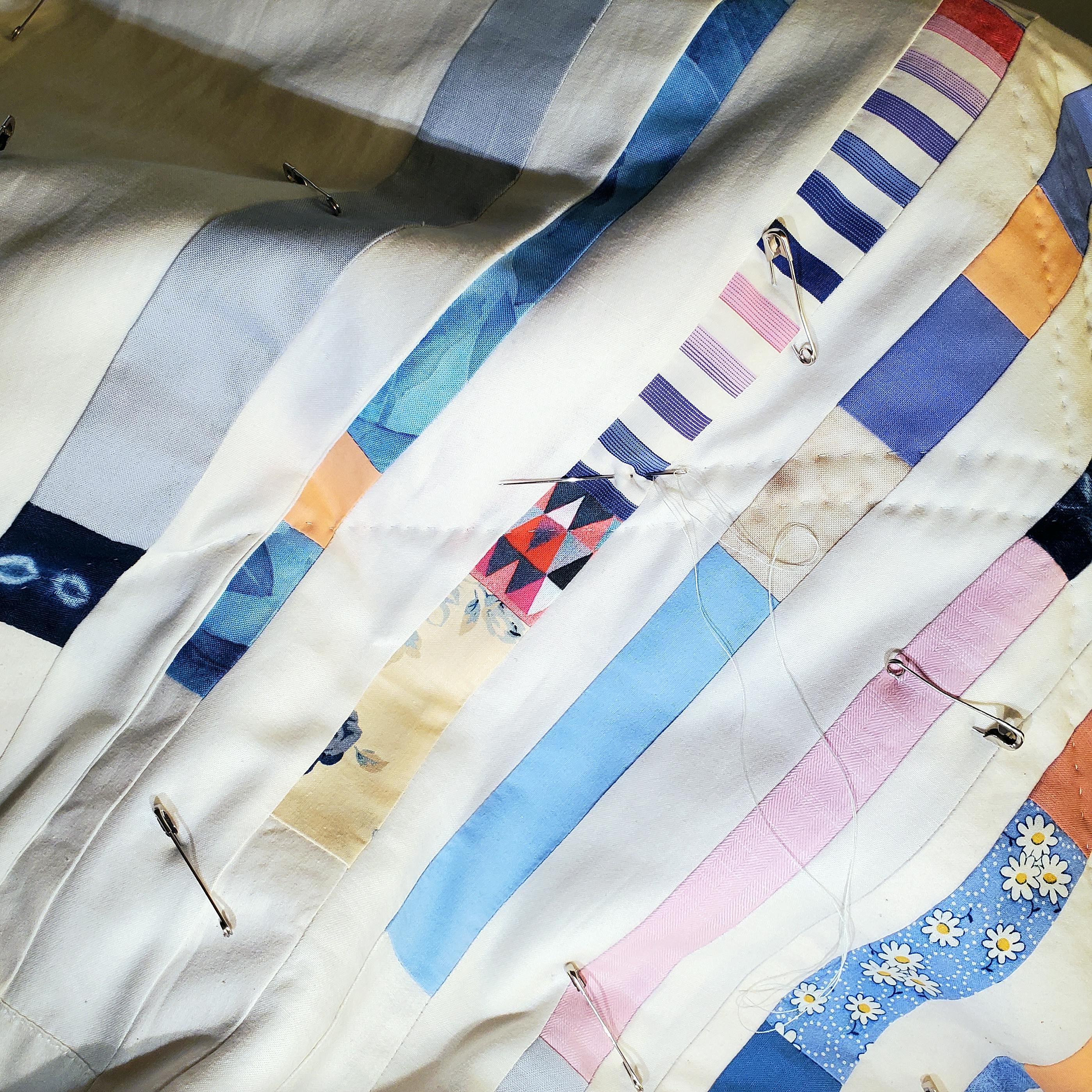 Close up photo of the patchwork quilt in progress, showing safety pins holding the quilt layers together, along with a needle and thread.