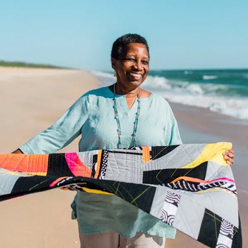 Photo of quilter Carole Lyles Shaw. She is on a beach, holding one of her own pieces.