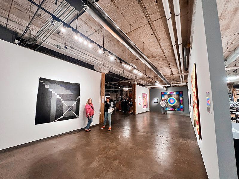 Photo of a gallery space with paintings and quilts hung on the walls. Two people are visible: Sarah in a black and white jacket, and Lela in a pink shirt.