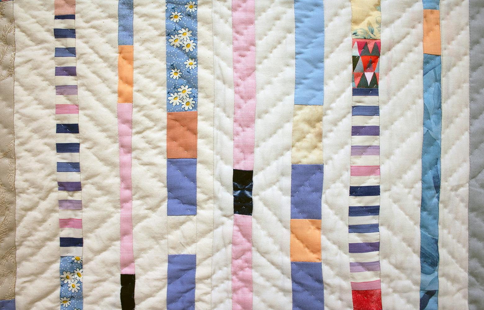 Detail photo of a patchwork quilt composed of pink, blue, and gray vertical stripes on a field of off-white. Diagonal lines of hand stitching are visible.