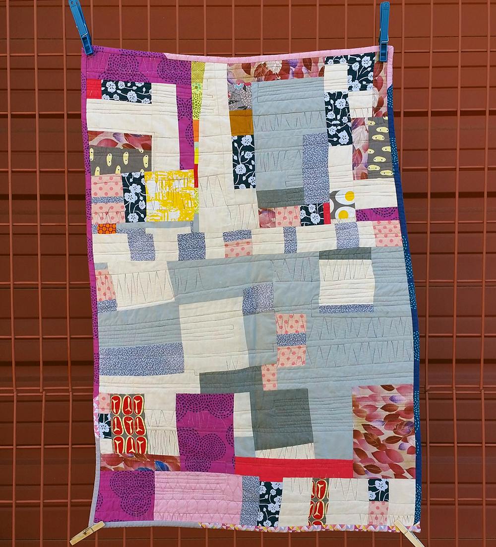 Photo of an improvised patchwork quilt clipped to a dark red wire gate in front of a garage door. The quilt contains fabrics in gray, off-white, pink, purple, yellow, and blue.