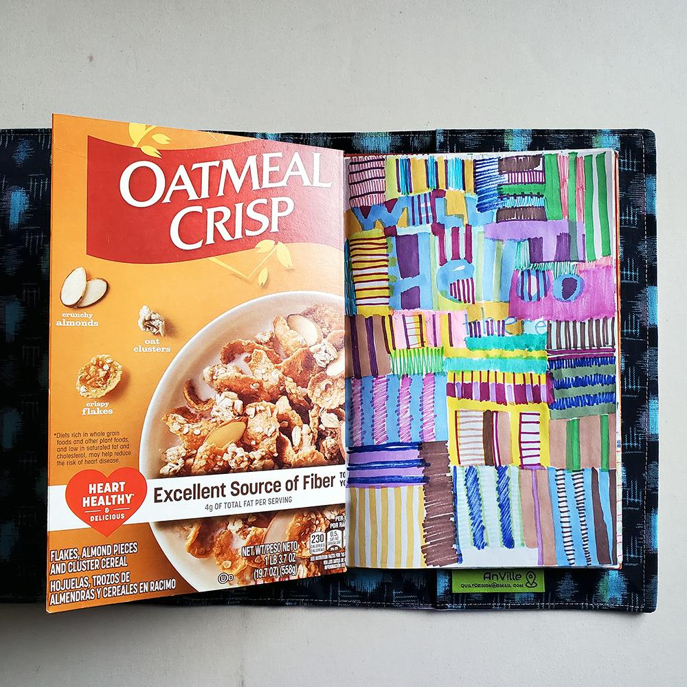 Photo of a homemade sketchbook in a fabric cover. The left page is a piece of cardboard cut from a cereal box. The right page is full of drawn stripes and squiggles.