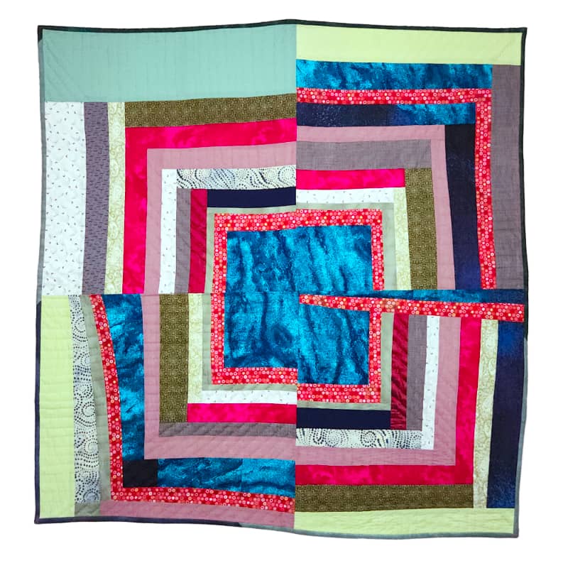 An improvised patchwork quilt, titled Wisconsin Baby, created by Sarah Atlee. The overall design is a central square of bright blue, interrupted by a red stripe. The colors of the surrounding fabric strips are red, pink, green, gray, and dark blue.